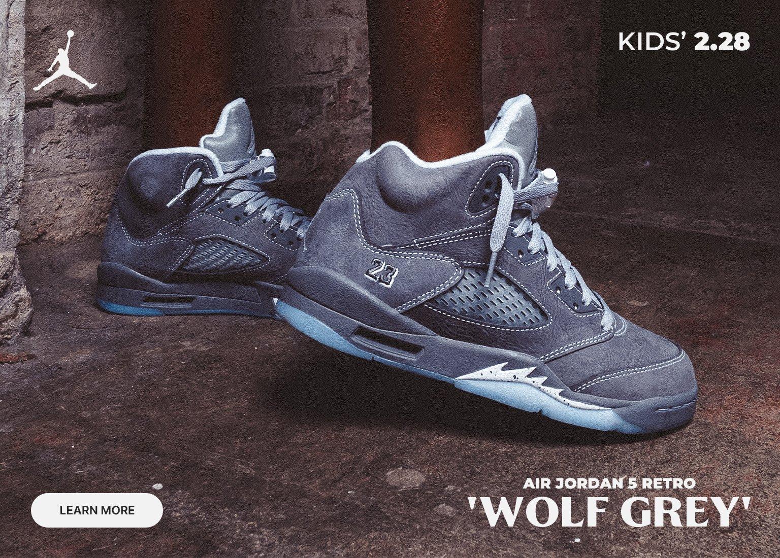 An advertisement for Jordan 5 Retro Wolf Grey sneakers featuring three different sizes of the grey suede shoes displayed in a row. The background is a dark, industrial floor and wall. Text includes the Jordan logo, the shoe name, LAUNCHES 02.28, and a LEARN MORE button.