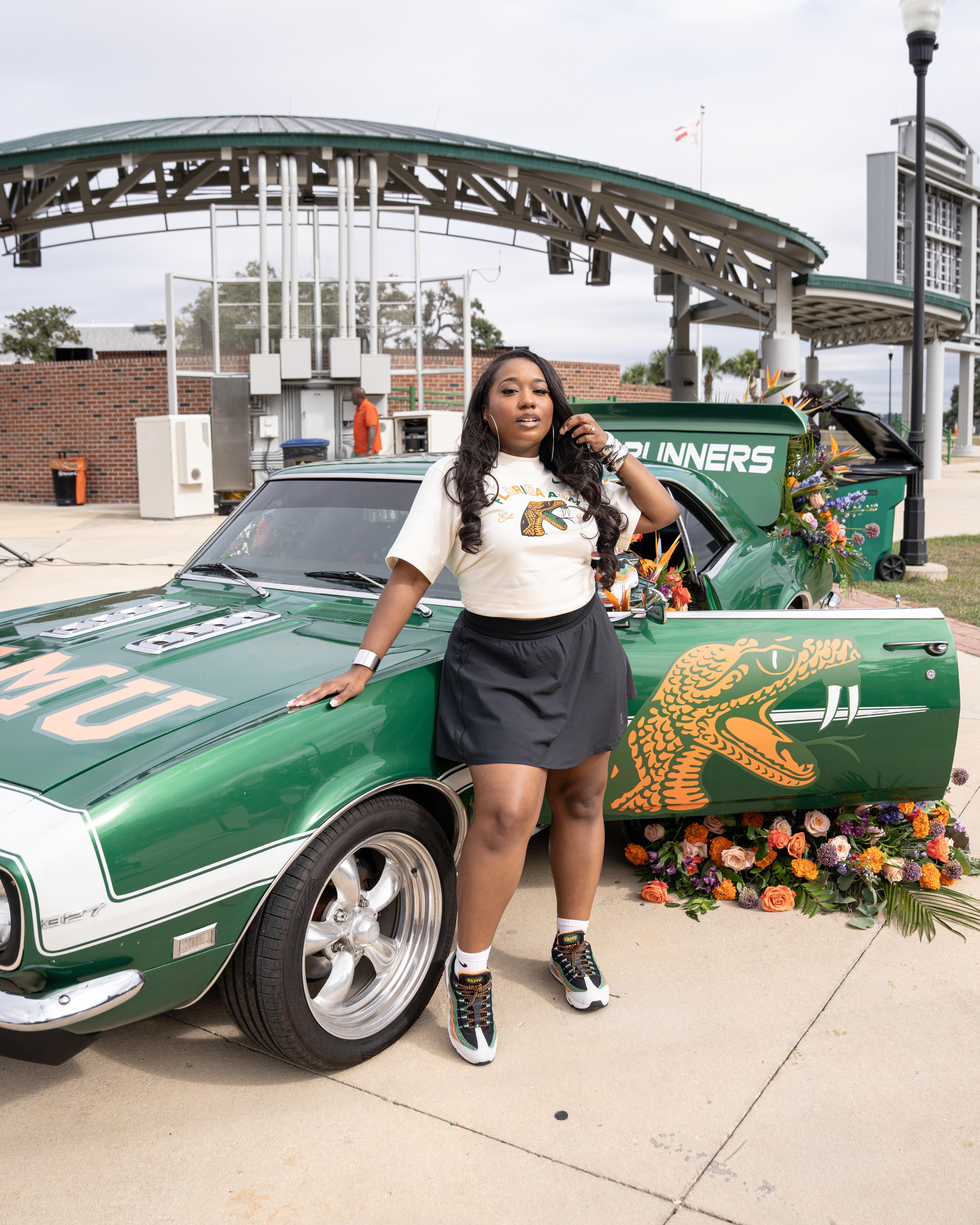 A stylish woman stands next to a vintage green muscle car, which is decorated with flowers and features the FAMU snake mascot design on the door. She is wearing a cream-colored t-shirt with the snake design and a black skirt.