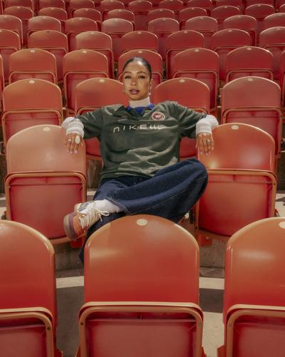 Person seated among orange stadium seats wearing a green Nike jersey, blue jeans and multicolor sneakers.