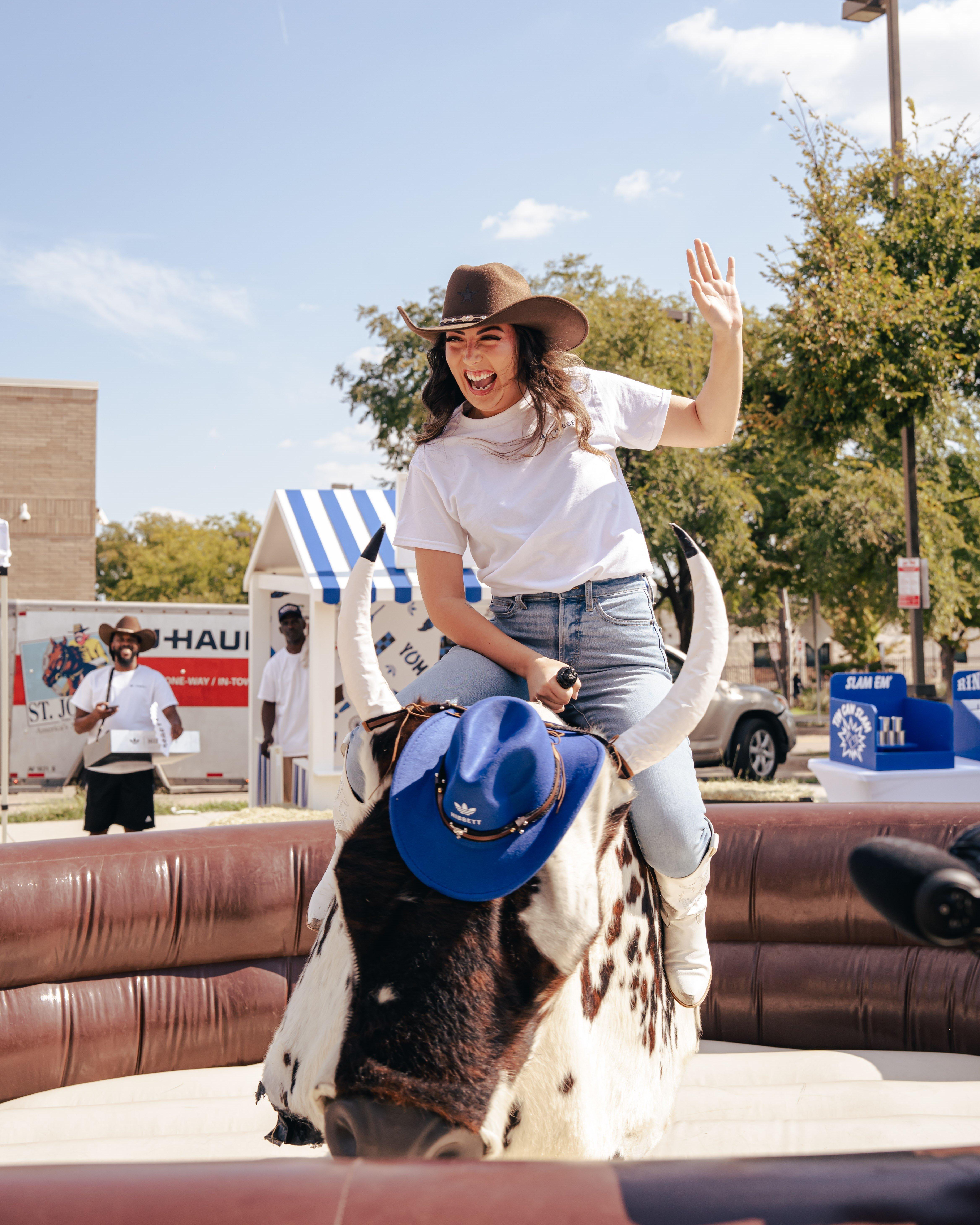 A woman in a white t-shirt, jeans, and cowboy boots rides an inflatable mechanical bull, smiling and waving. The bull has a blue cowboy hat on its head. In the background, other event attendees and a white tent are visible.