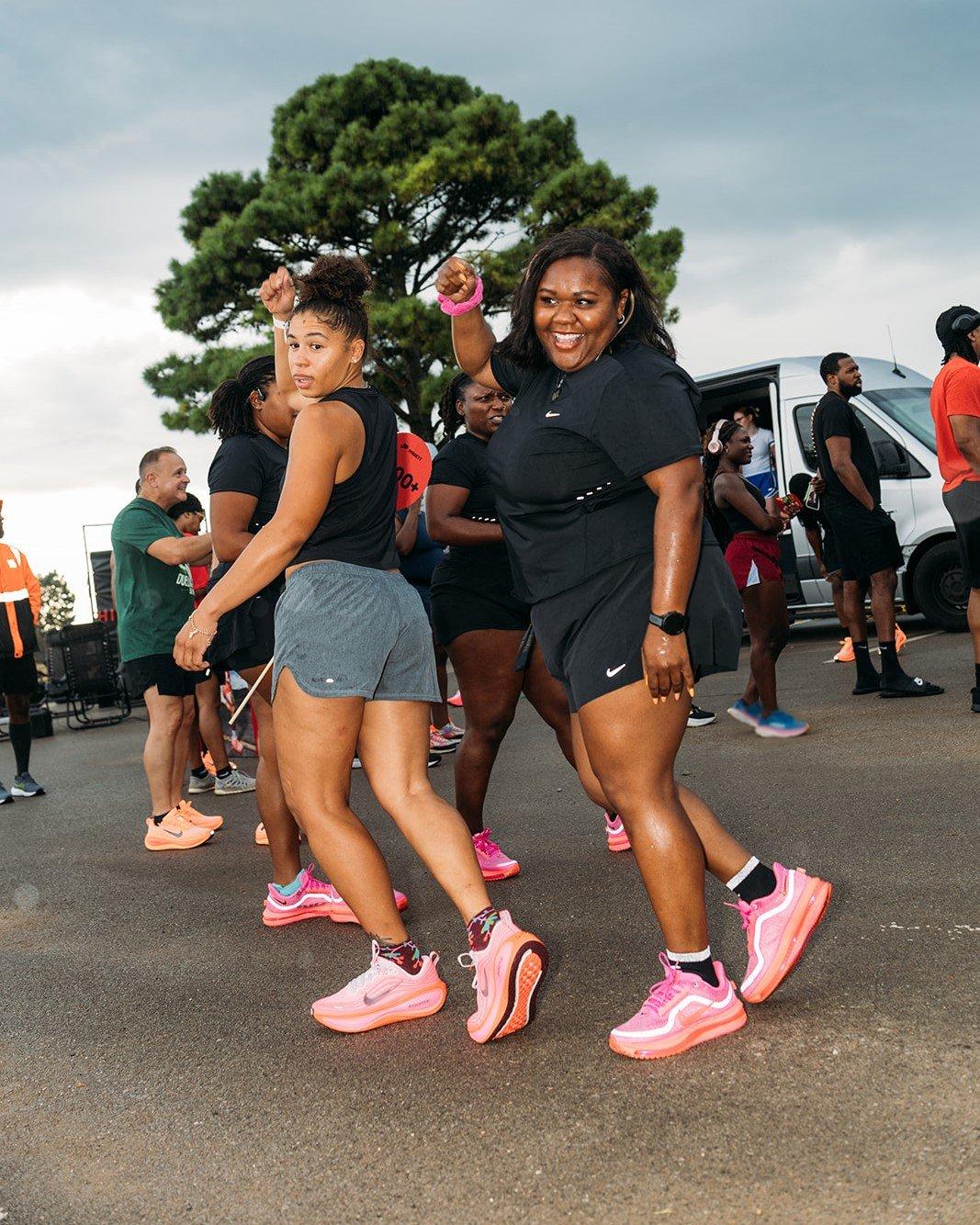Two women are smiling and dancing on a paved surface. Both are wearing running clothes and brightly colored pink running shoes. Other people are visible in the background, including a white van. The ground is wet.