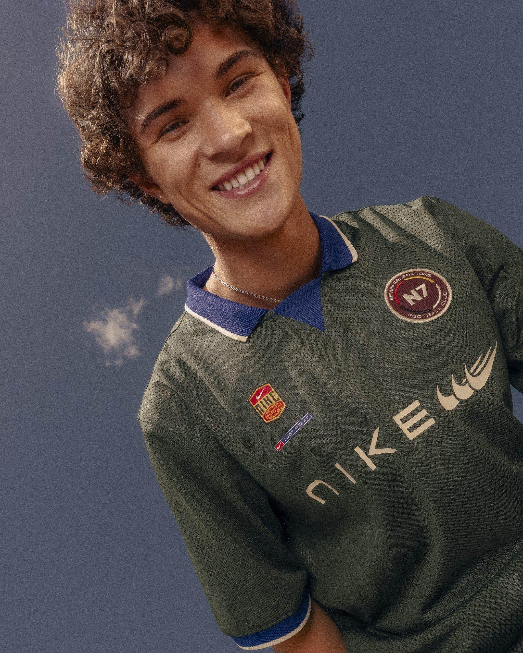 A smiling young man looking down at the camera against a blue sky, wearing a dark green textured Nike N7 soccer jersey. The mesh jersey features a blue collar, a circular "Seven Generations Nike N7 Football Club" crest on the left chest, a vintage-style Nike logo badge on the right, and gold "NIKE" lettering printed across the front.