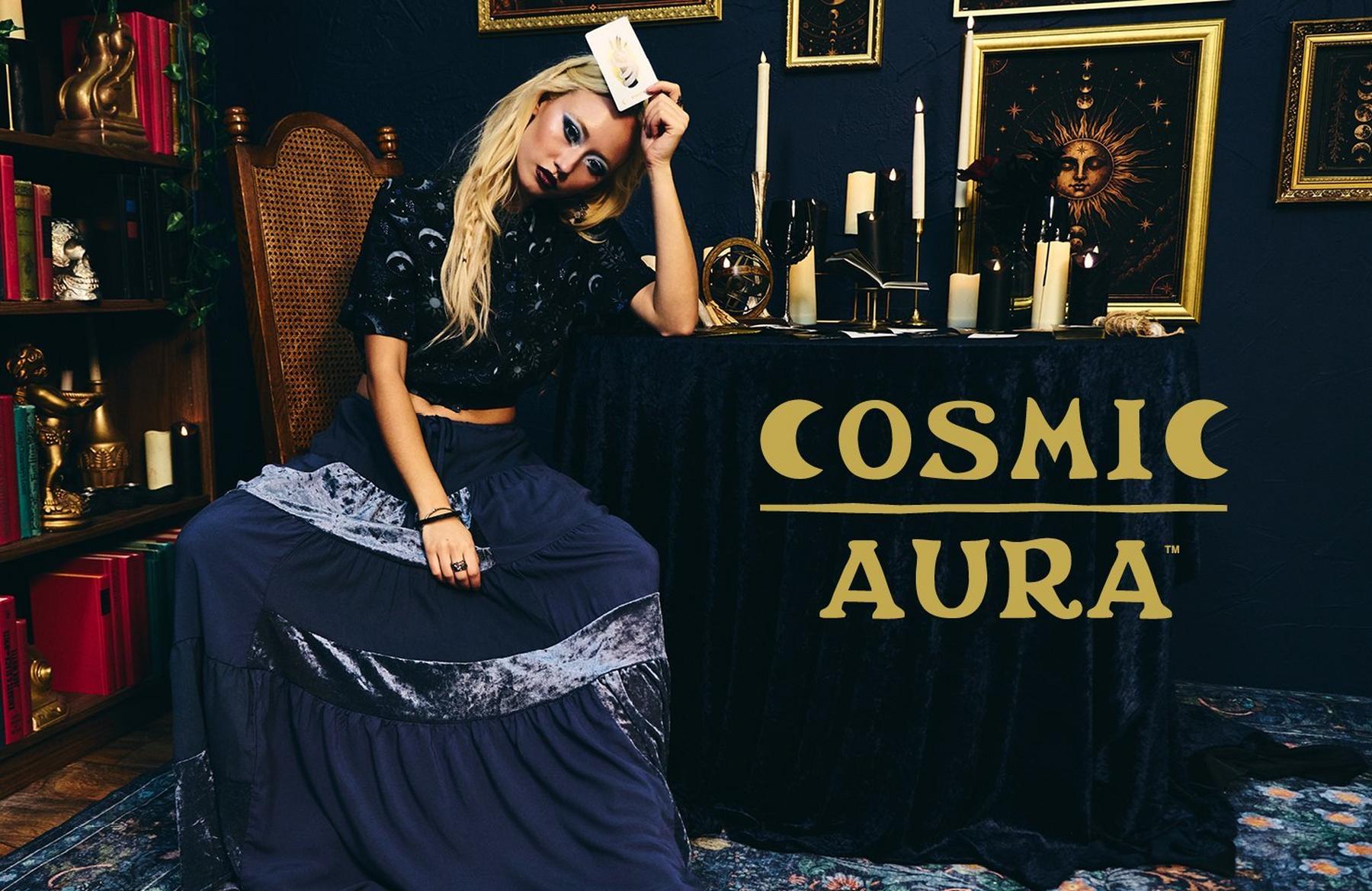 Model in dark celestial outfit holding a tarot card beside a table with “COSMIC AURA” text. Shop Now.