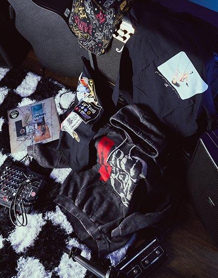 Assorted graphic hoodies, T-shirts, patches, and stickers laid out on a rug. Shop Now.