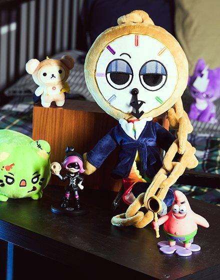 Plush clock-faced doll in a suit, surrounded by small toy figures on a table. Shop Now.