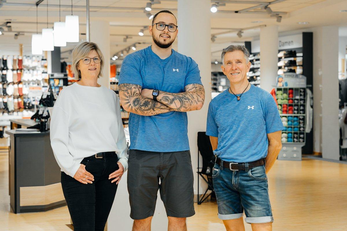 Drei Menschen stehen in einem Sportladen. Die linke Person trägt eine Brille, ein weißes Oberteil und eine schwarze Hose, die Person in der Mitte hat dunkle Haare, eine Brille, einen Bart und trägt ein blaues T-Shirt und kurze Hose, die dritte Person von links hat graue Haare,  trägt ein blaues T-Shirt und eine kurze Hose. 