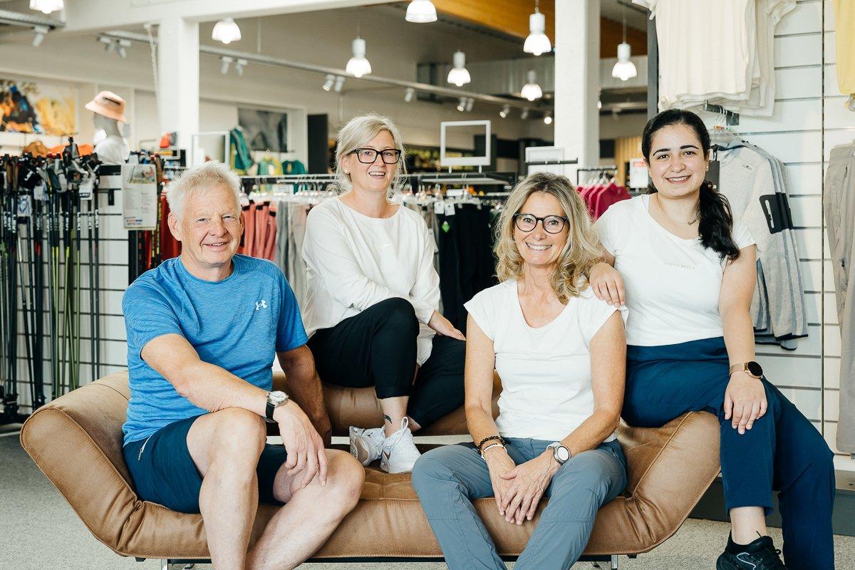 Vier Menschen sitzen gemeinsam auf einem braunen Sofa in einem Geschäft. Die Person links trägt ein blaues T-Shirt und eine blaue, kurze Hose und hat kurze, graue Haare, die Person rechts daneben trägt eine schwarze Brille, ein weißes T-Shirt, eine schwarze lange Hose und hat blonde Haare. Die dritte Person von links hat lange blonde Haare und eine schwarze runde Brille, sie trägt ein weißes T-Shirt und eine graue, lange Hose. Die Person rechts hat schwarze Haare und trägt ein weißes T-Shirt und eine lange, blaue Hose.