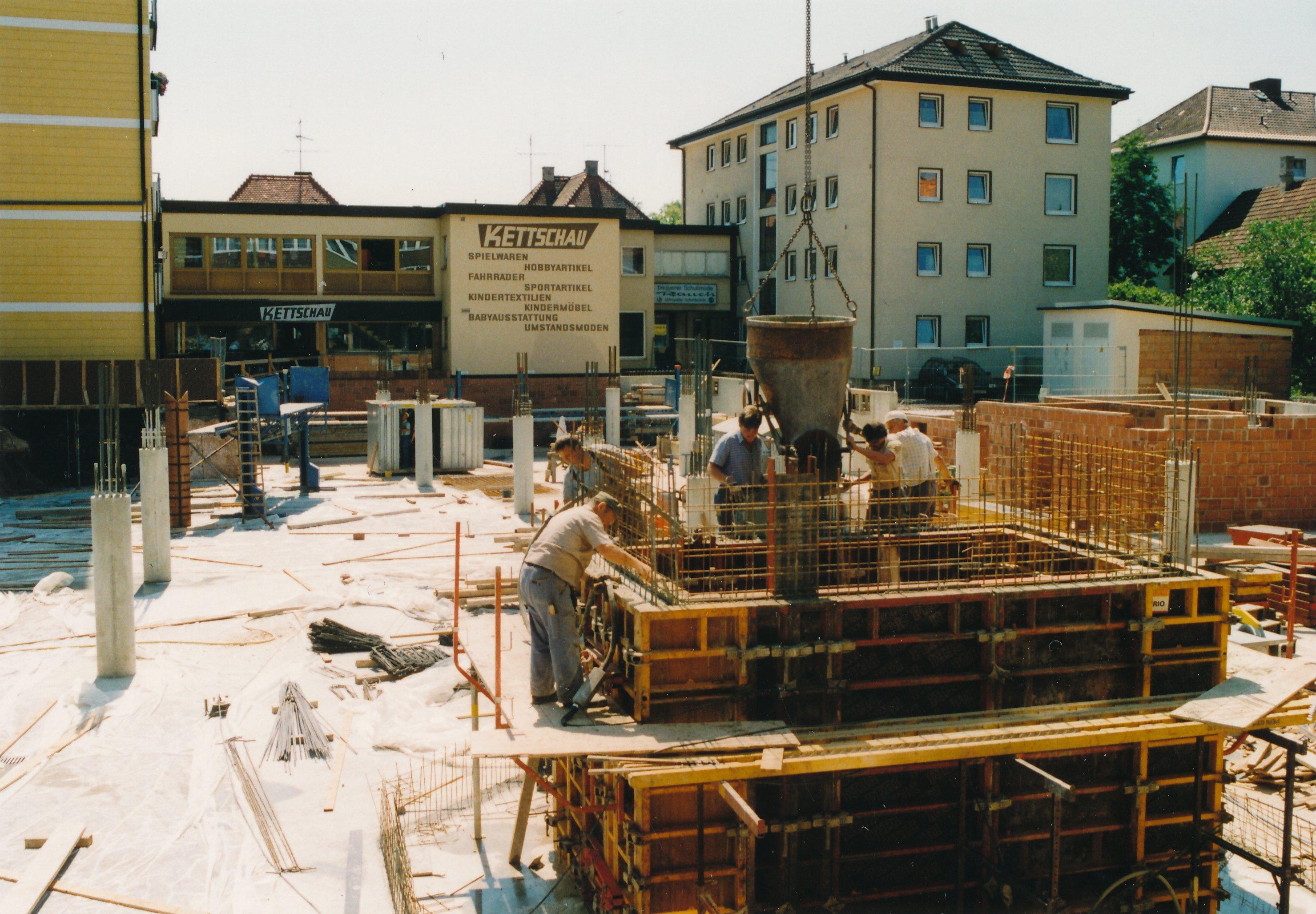 Eine Baustelle: Hinten in der Mitte steht eine Hauswand mit dem Logo "Kettschau" dran, daneben ein weißes Wohnhaus, im Vordergrund wird auf der Baustelle gebaut. 