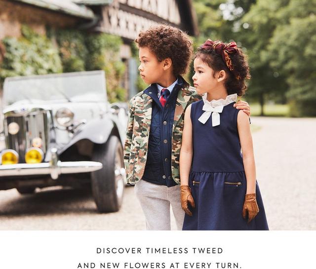Discover timeless tweed and new flowers at every turn. Image: Boy wearing camo jacket over quilted blue barn coat with gray joggers. His arm is around a girl wearing a blue dress with a white bow. Background of old car and scenic barn.