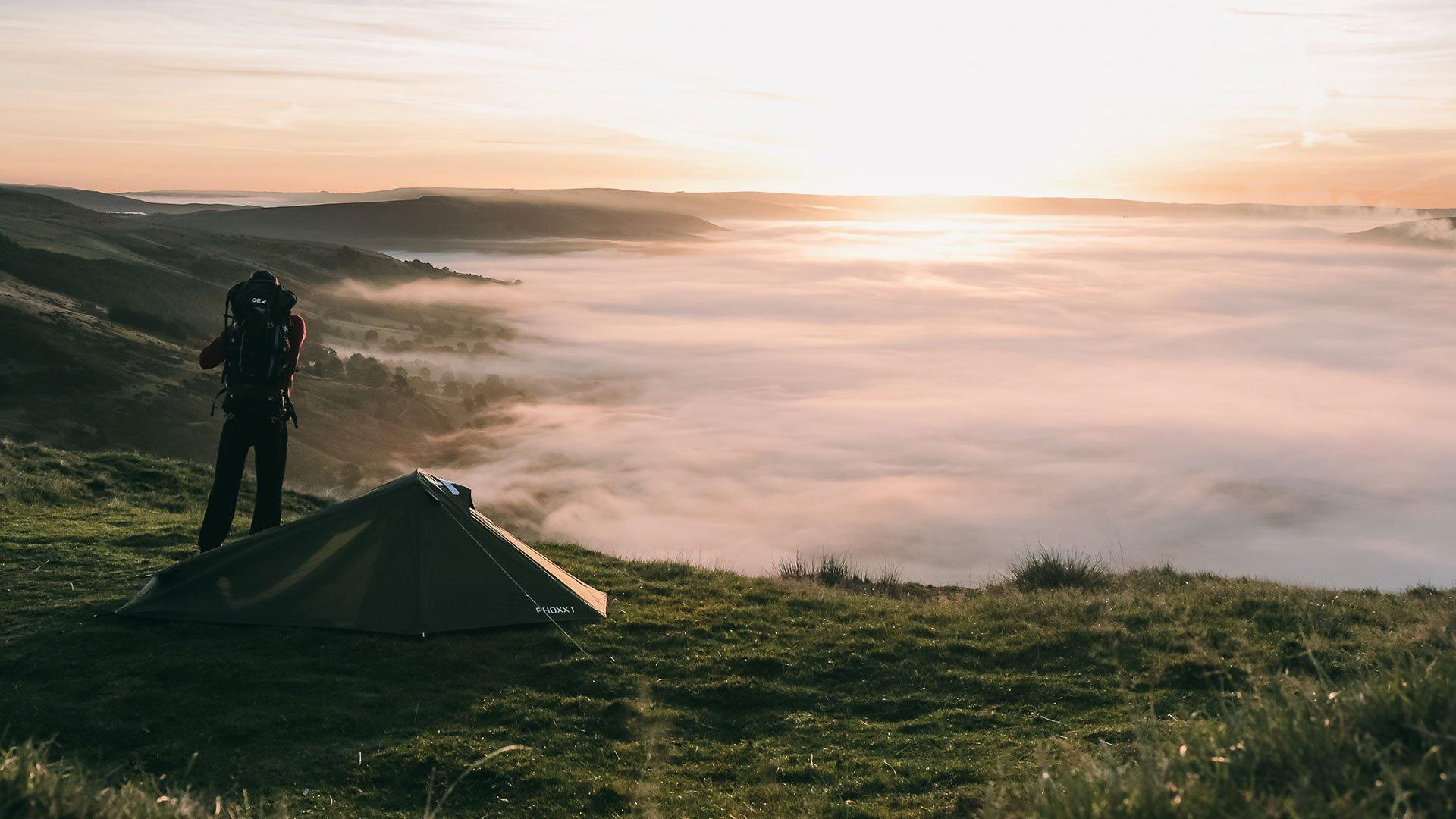 An image of man looking over a mountain range with a backpacking tent in the foreground at sunset