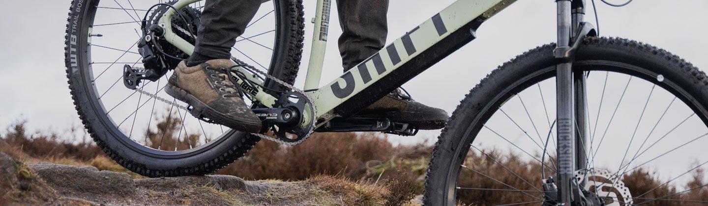 Closeup of a person riding a mountain bike on a muddy path with the focus on the shoe and pedals