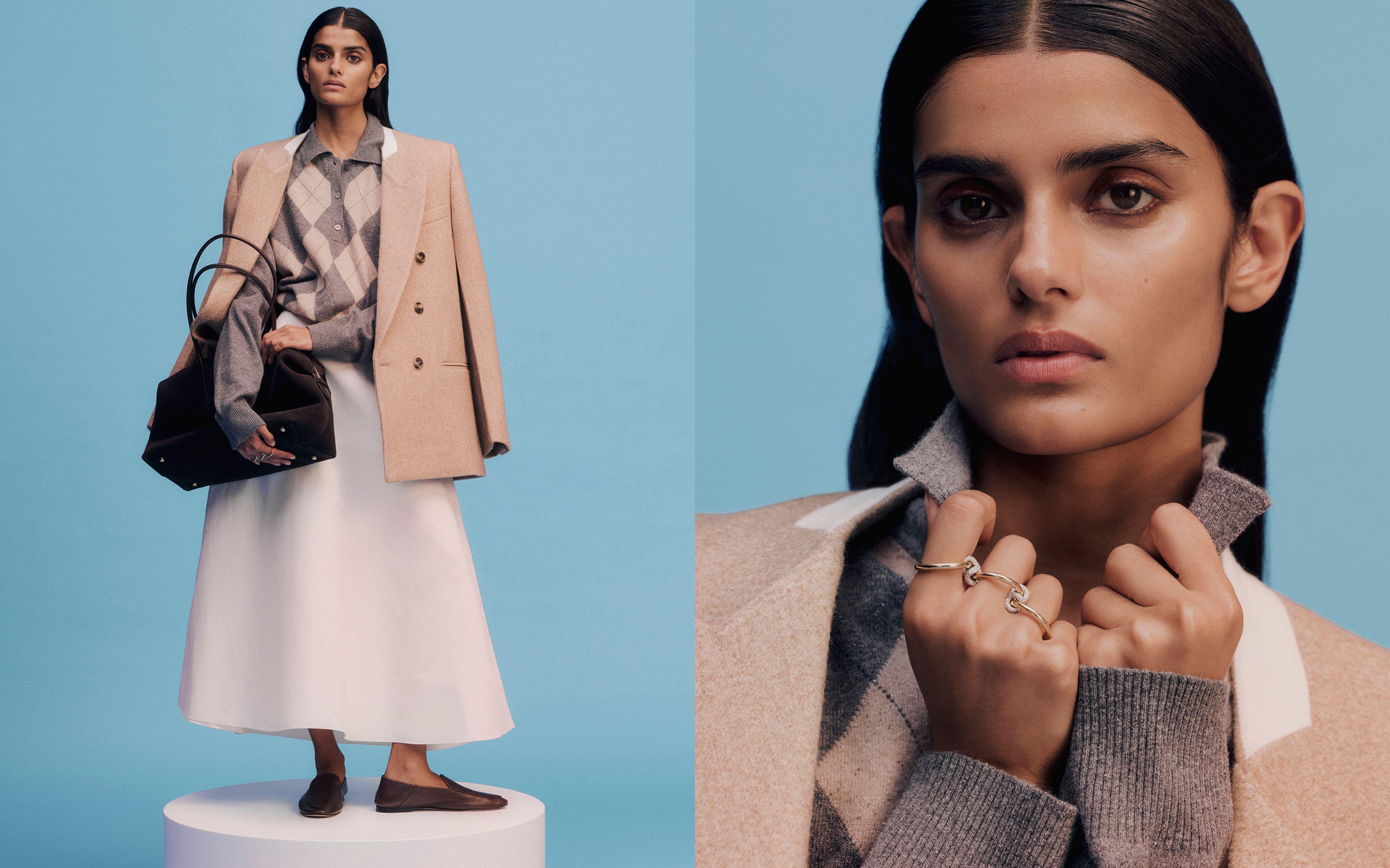 Model holding a black leather tote bag, wearing a white A-line skirt, leather flats, an argyle cardigan, neutral blazer and gold rings 