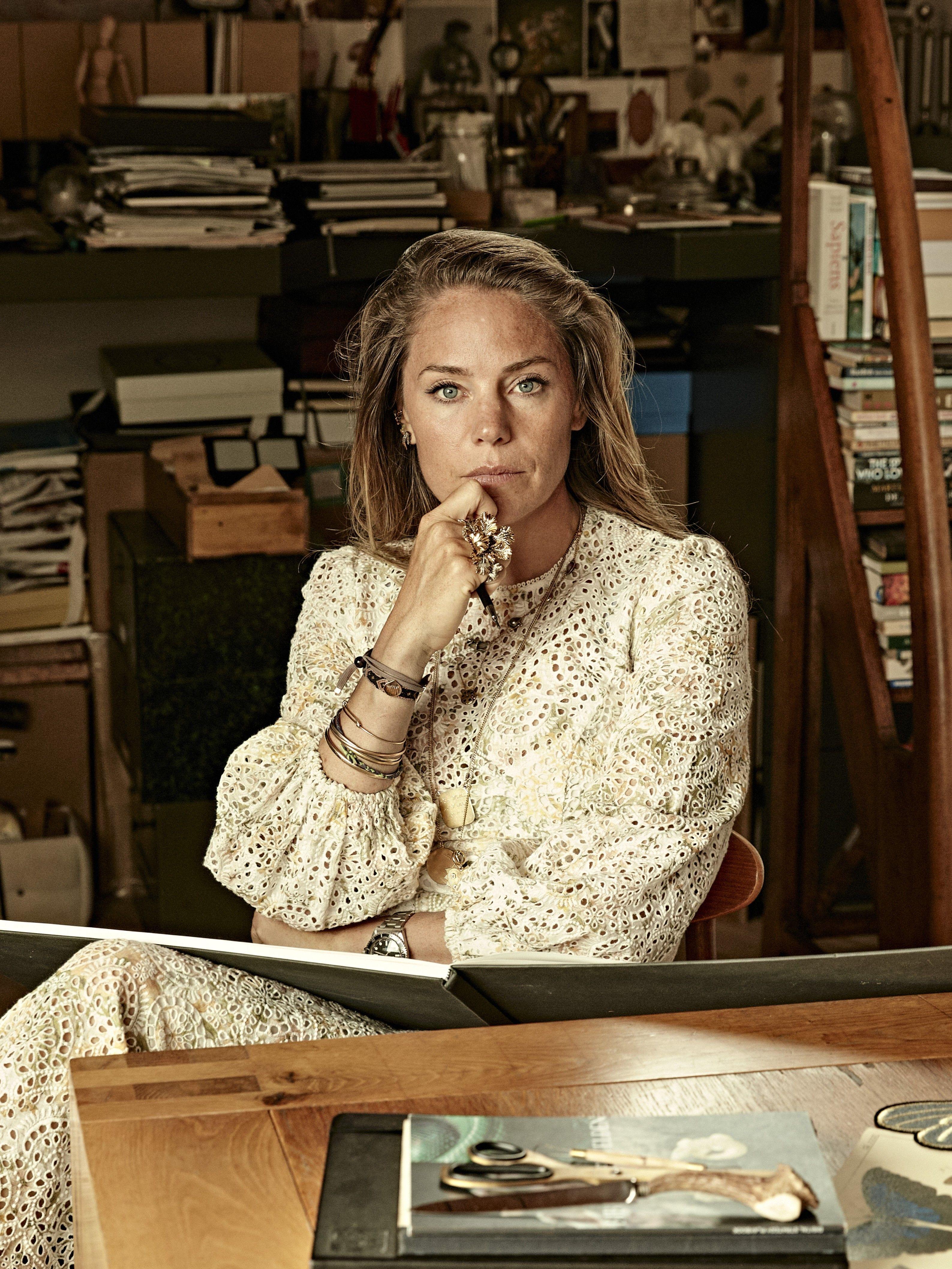 Bibi Van Der Velden sits in her studio. She wears a boho style lace eyelet dress, with many bracelets stacked up her arm. Her room is piled high with books and sketches, with interesting objects scattered around the surfaces. 