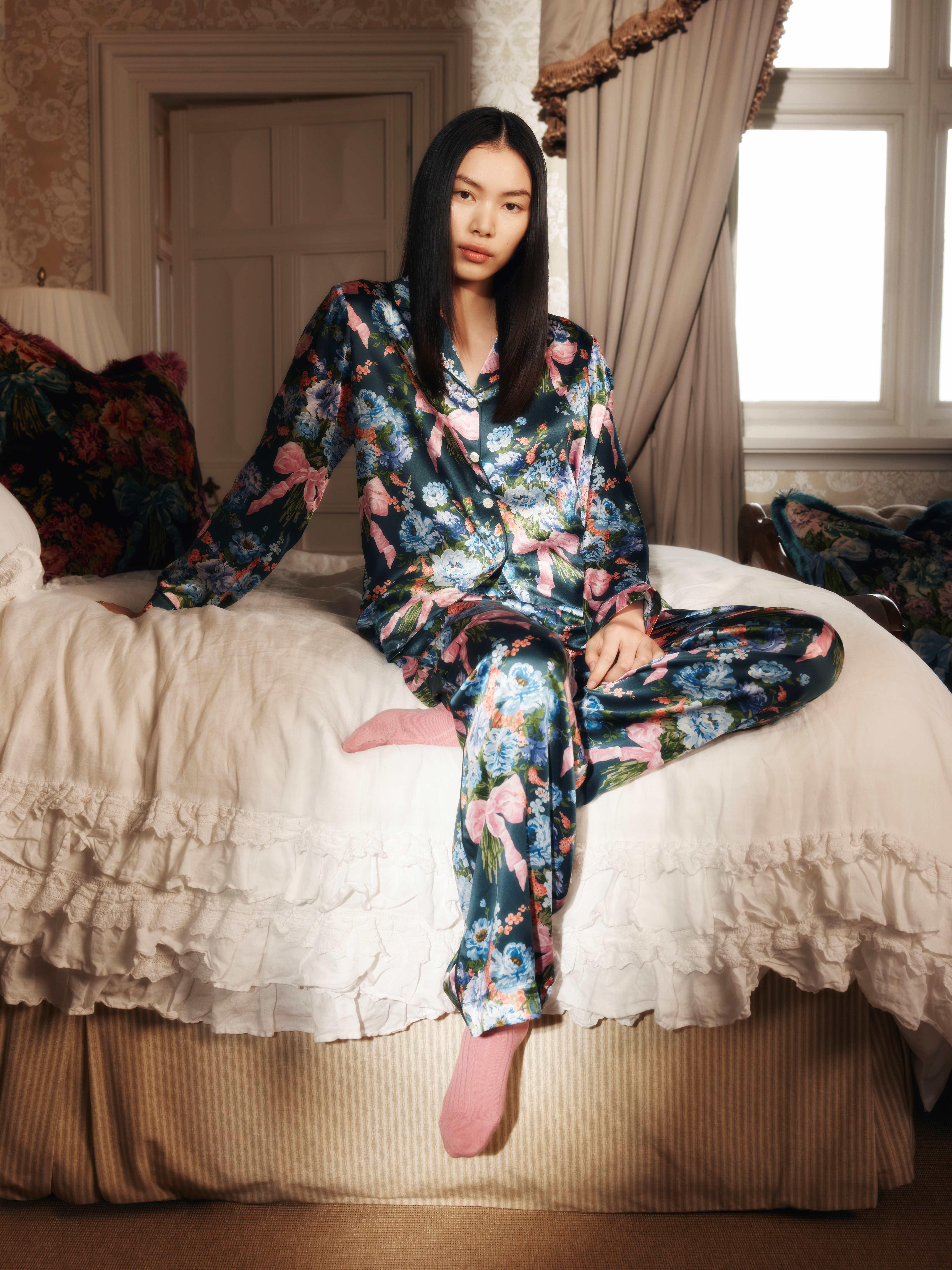 Model sitting on a bed wearing a silk Liberty Fabrics & Bridgerton pyjama set and pink socks 