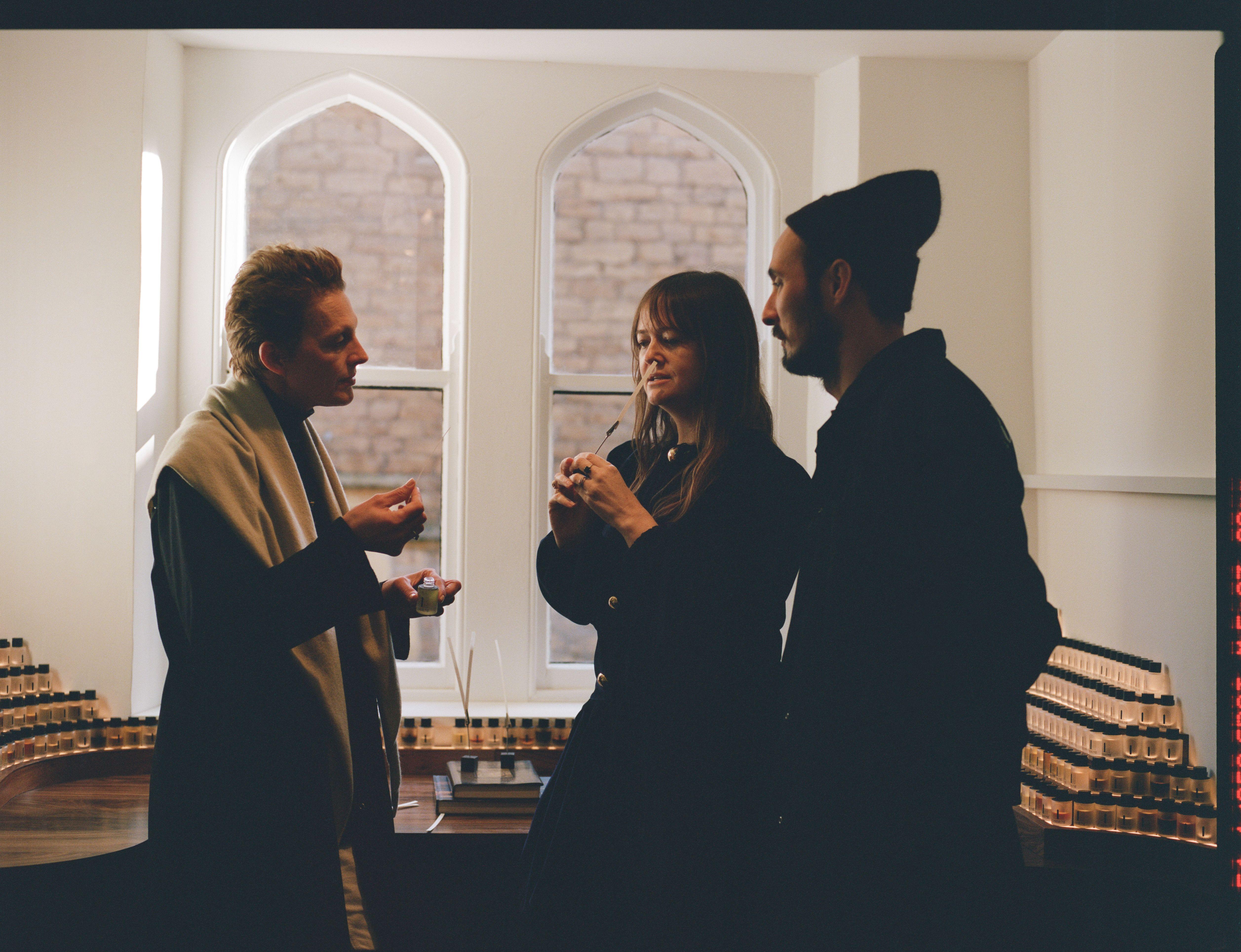 Three people stand in a low lit room, they are smelling fragrances on small paper tabs.