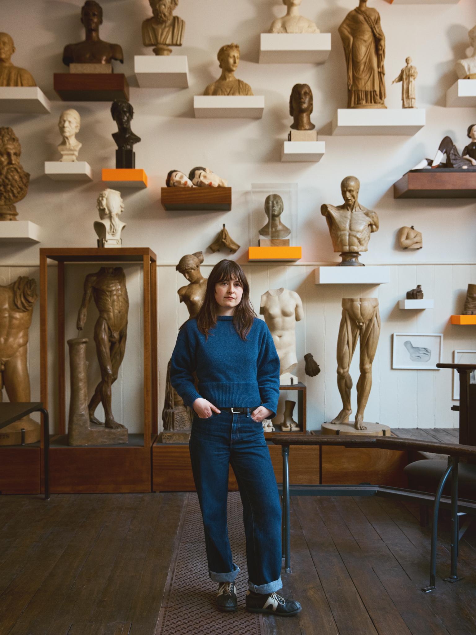 An artist wears blue overalls and stands in front of a wall of sculptures