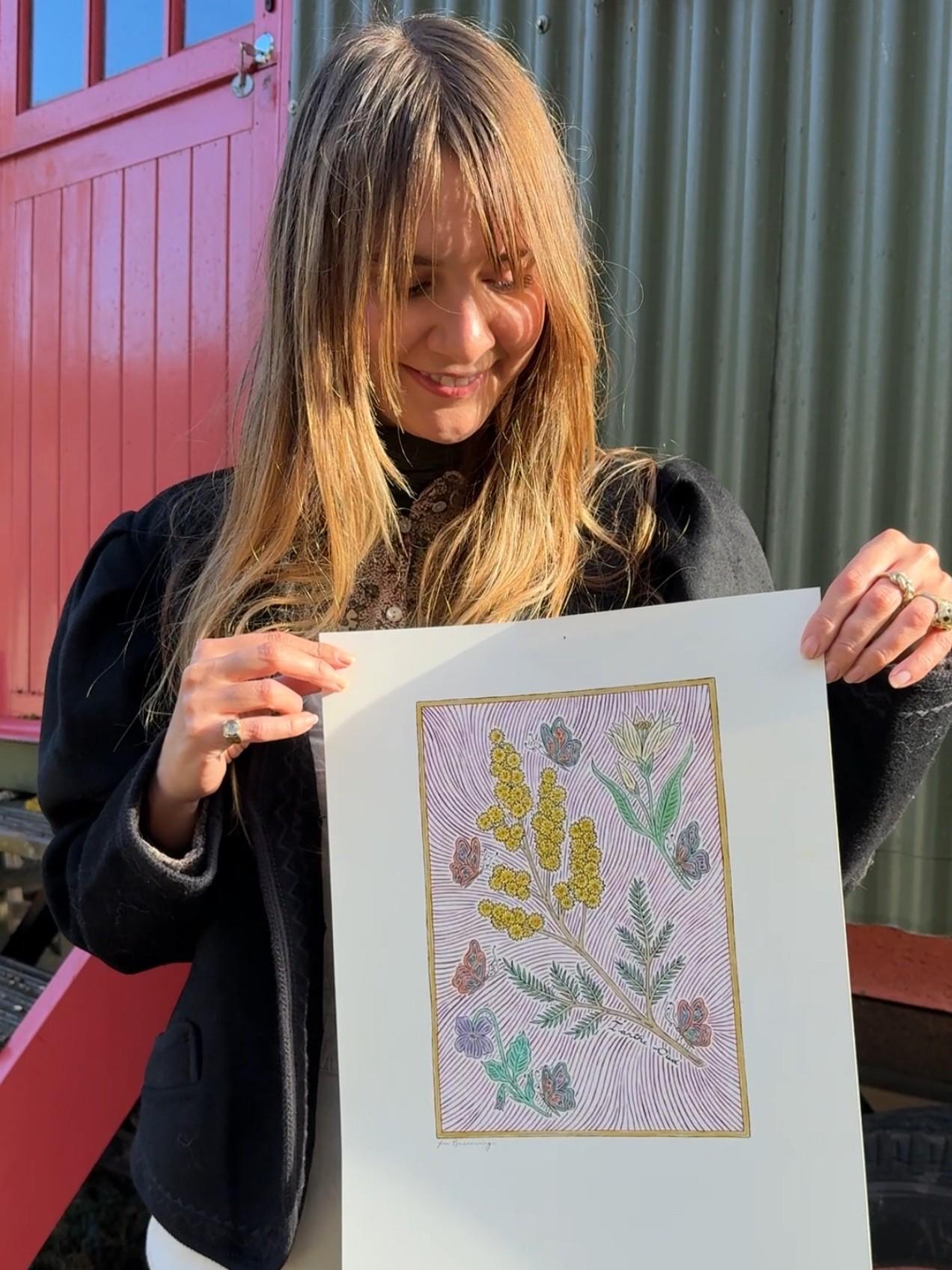 Fee Greening holds her artwork standing outside a red and green woodland cabin