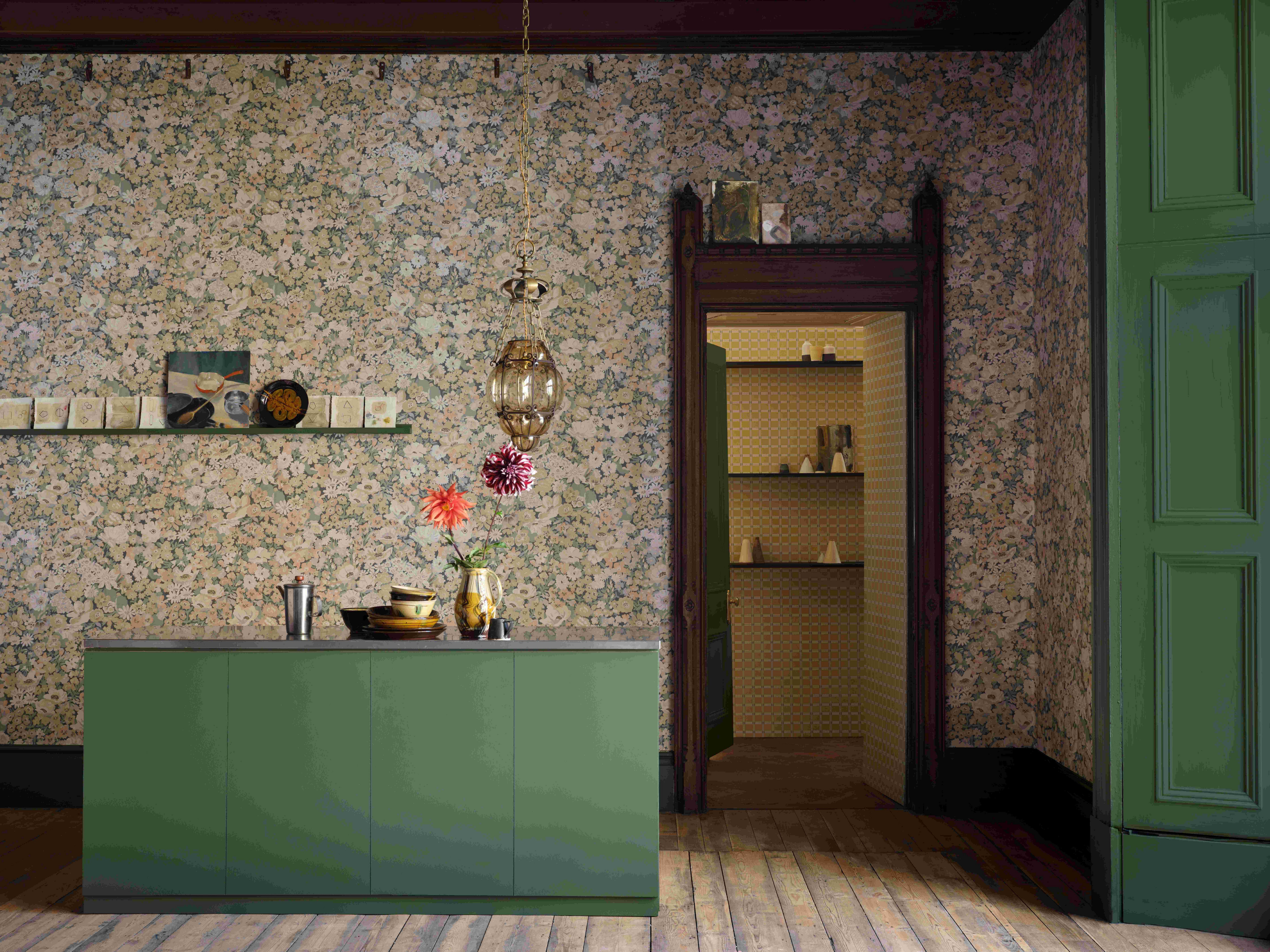 A kitchen with a wooden door frame, a green kitchen island and a decorated shelf, covered in Liberty Interiors Painted Thorpe in Lichen wallpaper