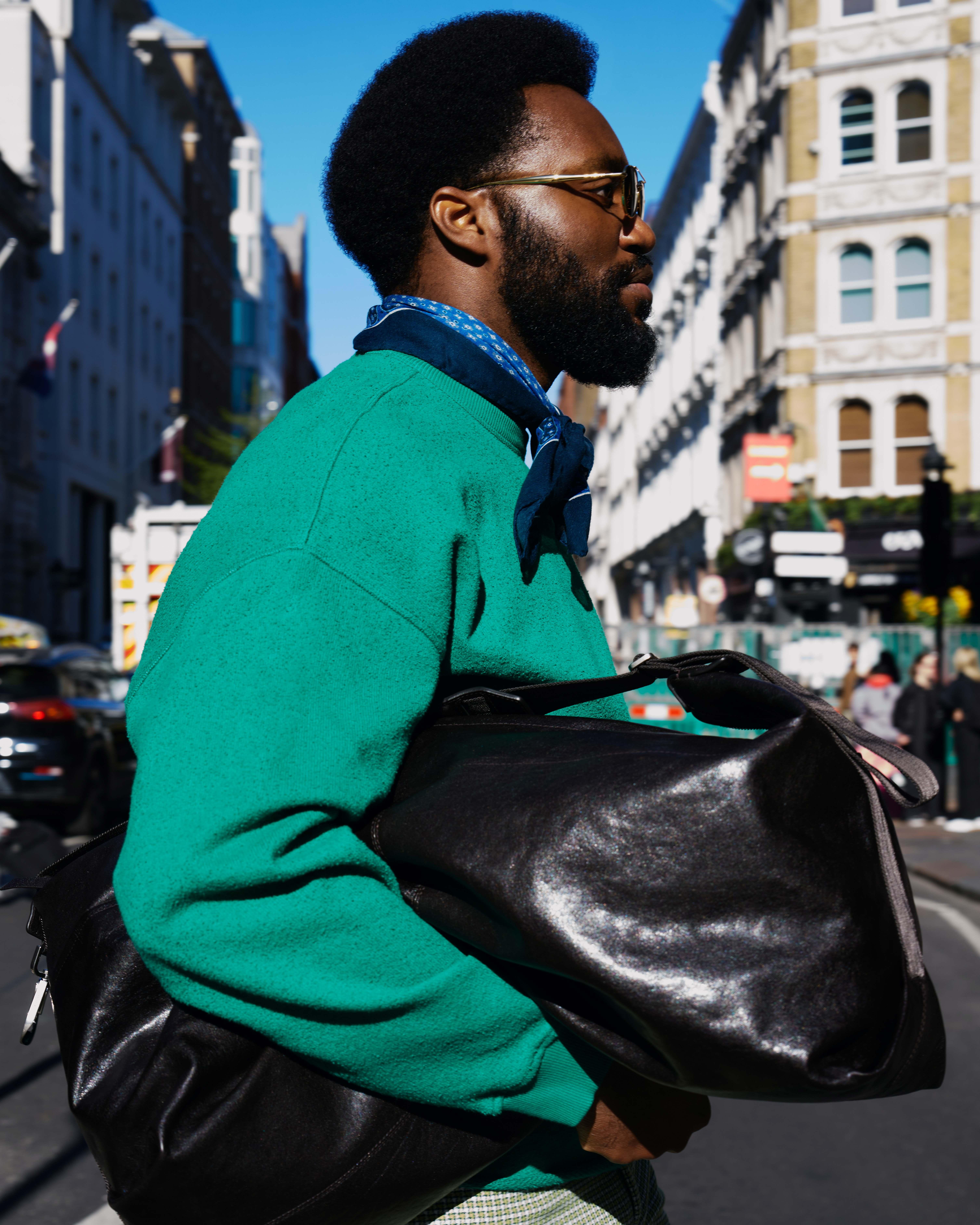 Okoya Obi crossing a road wearing a large black leather clutch, green jumper and glasses