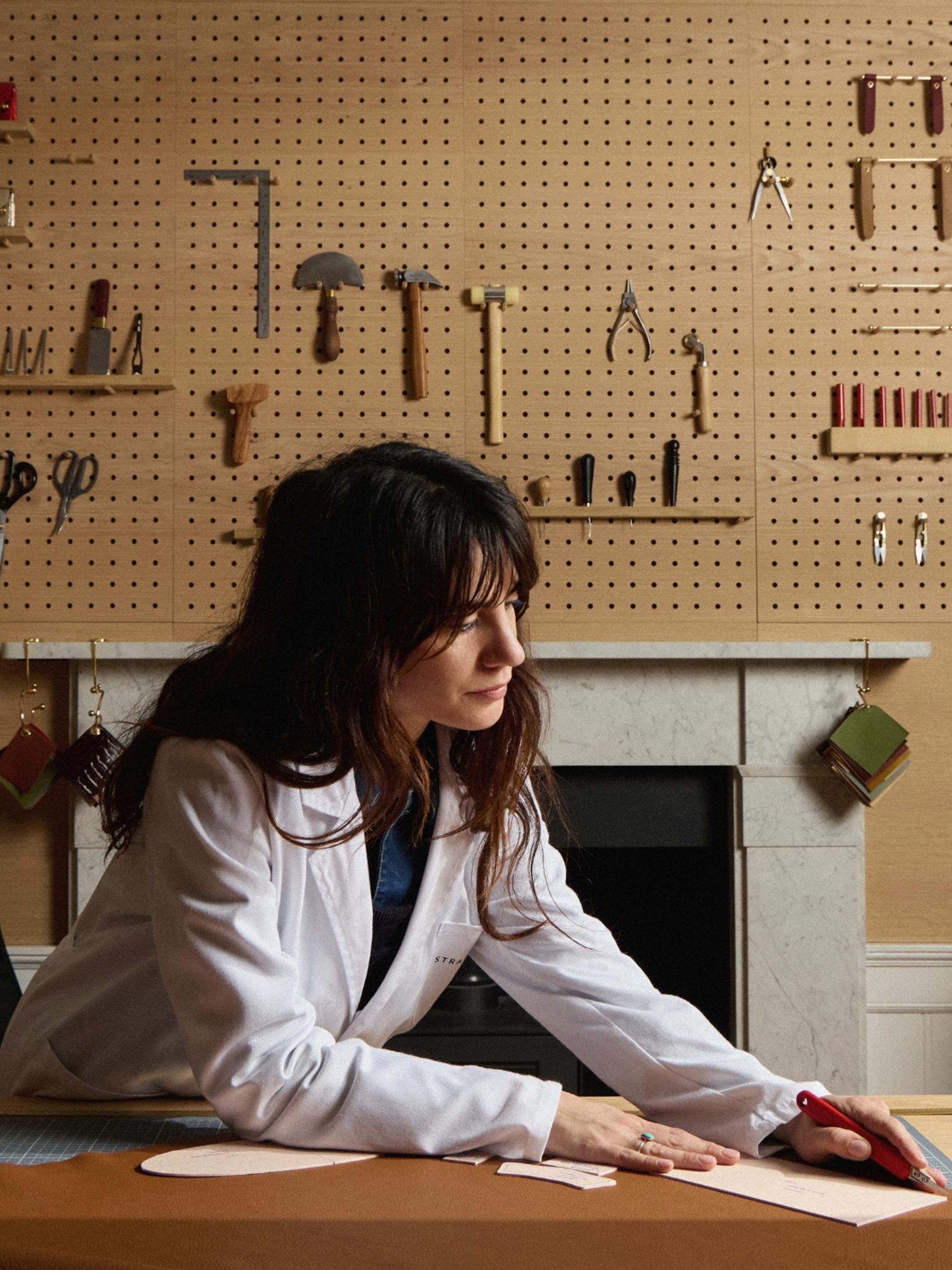 Head artisan Manon works on cutting leather from a larger piece. On the wall behind her are various hammers and tools of the leather working trade. 