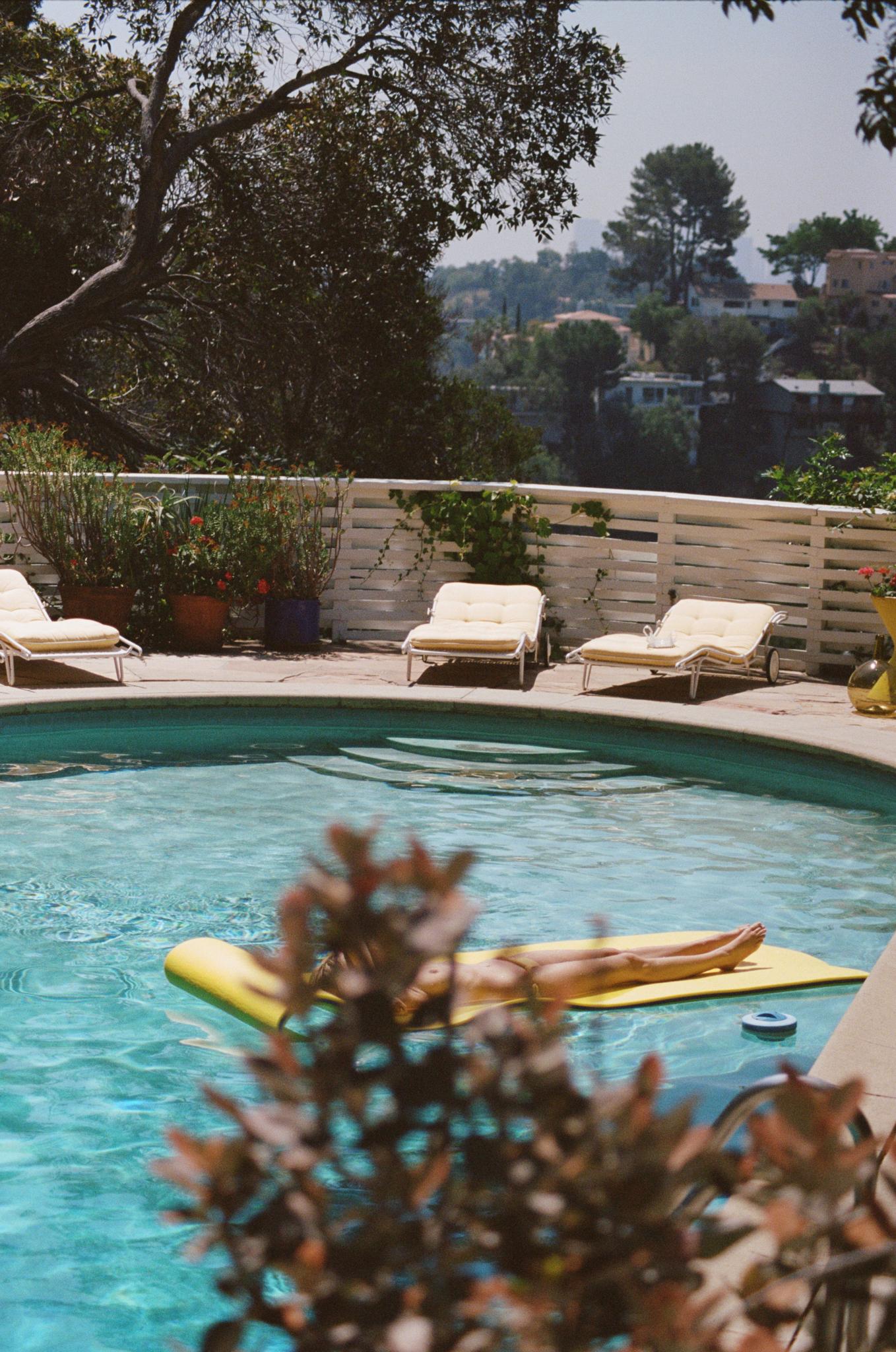 A pristine blue pool in the sunshine surrounded by trees and yellow sunloungers