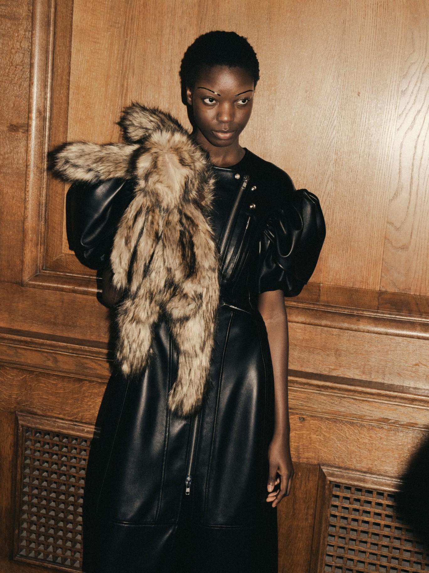 A model stands in front of a wood panel wall, wearing a puff sleeved leather dress and faux fur stole created by Simone Rocha