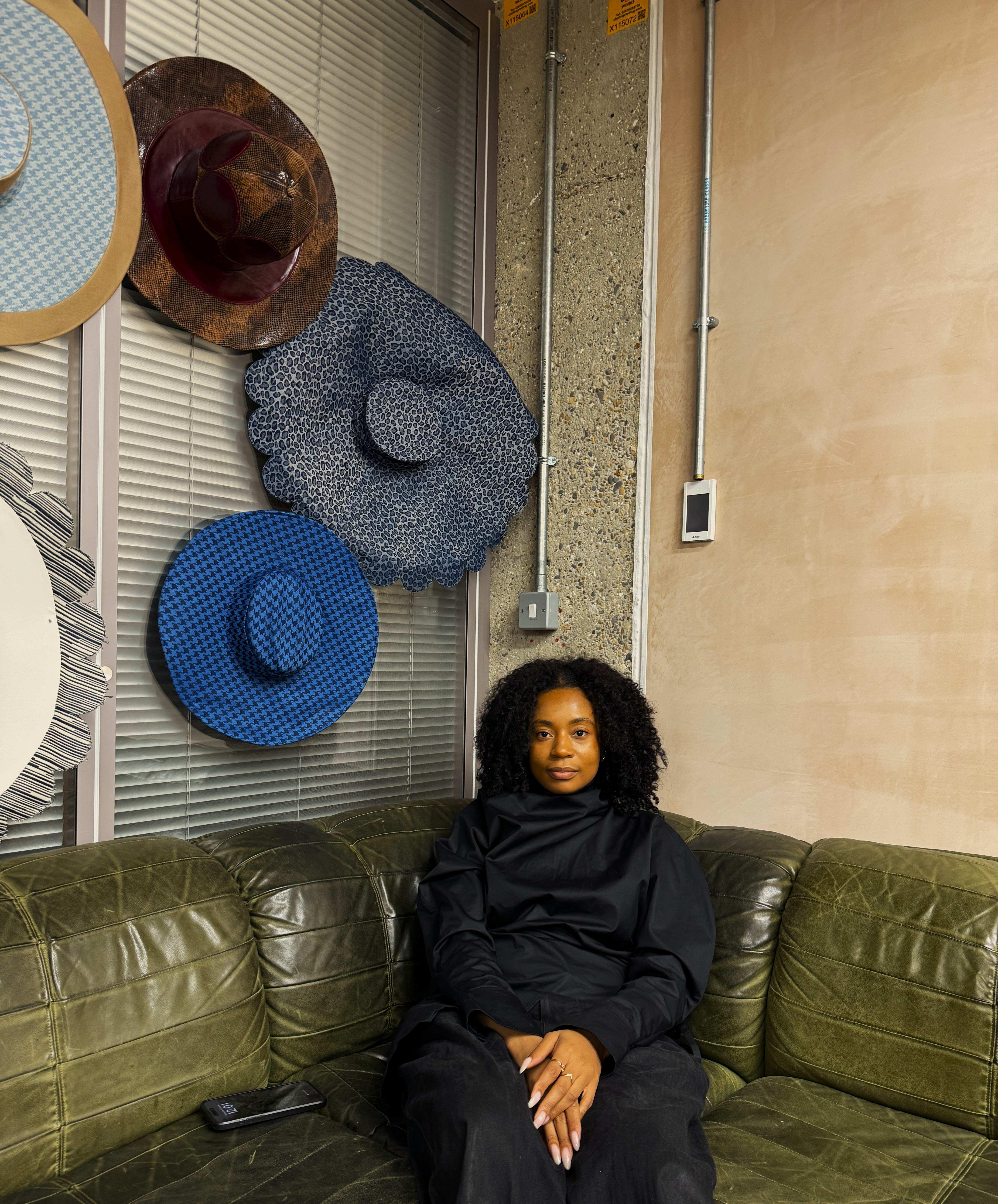 Tolu Coker sits on a green leather sofa, surrounded by hats she's designed 