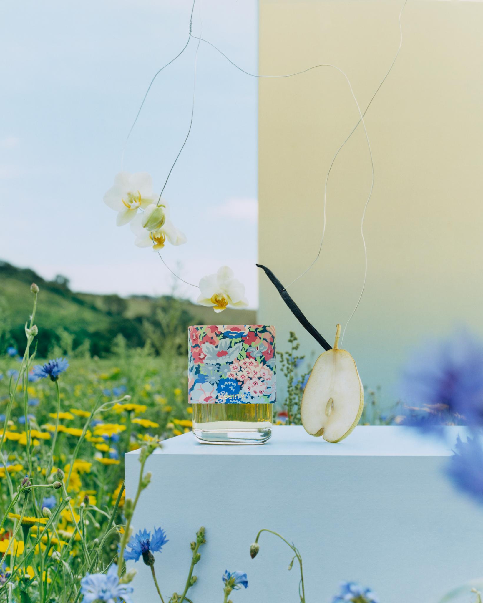 Liberty LBTY. Fragrance perfume bottle with floral cap on a pedestal in a summer meadow, surrounded by flowers and a sliced pear.