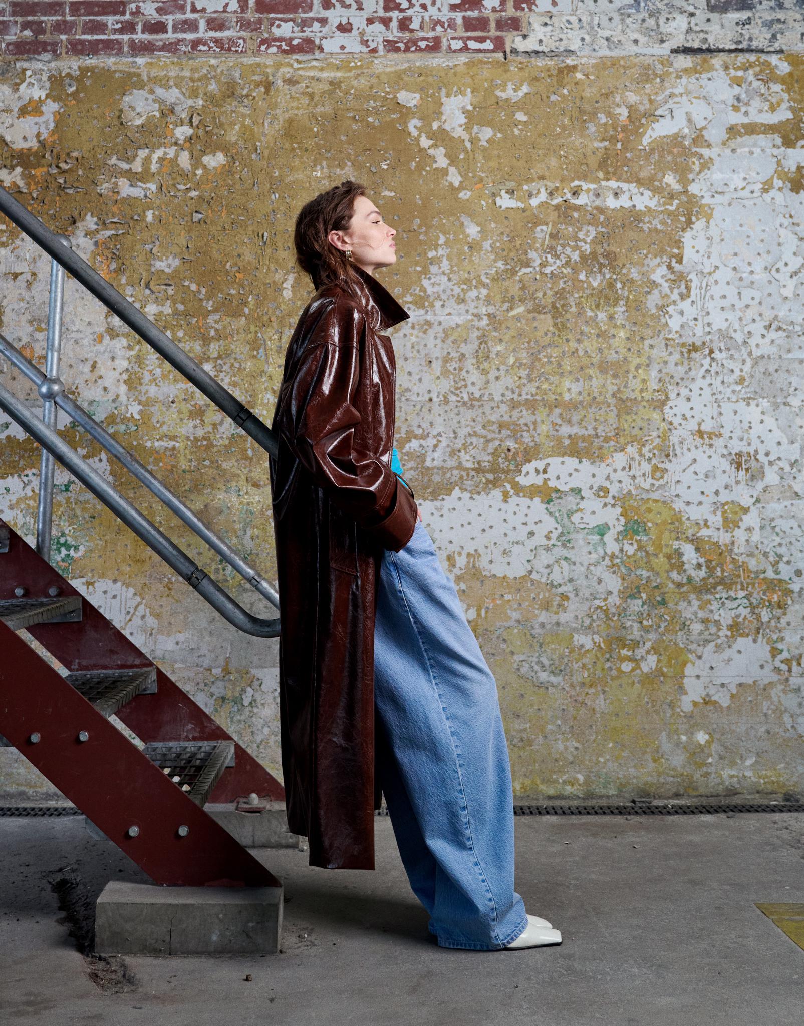 A woman wearing a shiny trench coat and baggy jeans from Topshop leans against a staircase in front of a distressed wall 