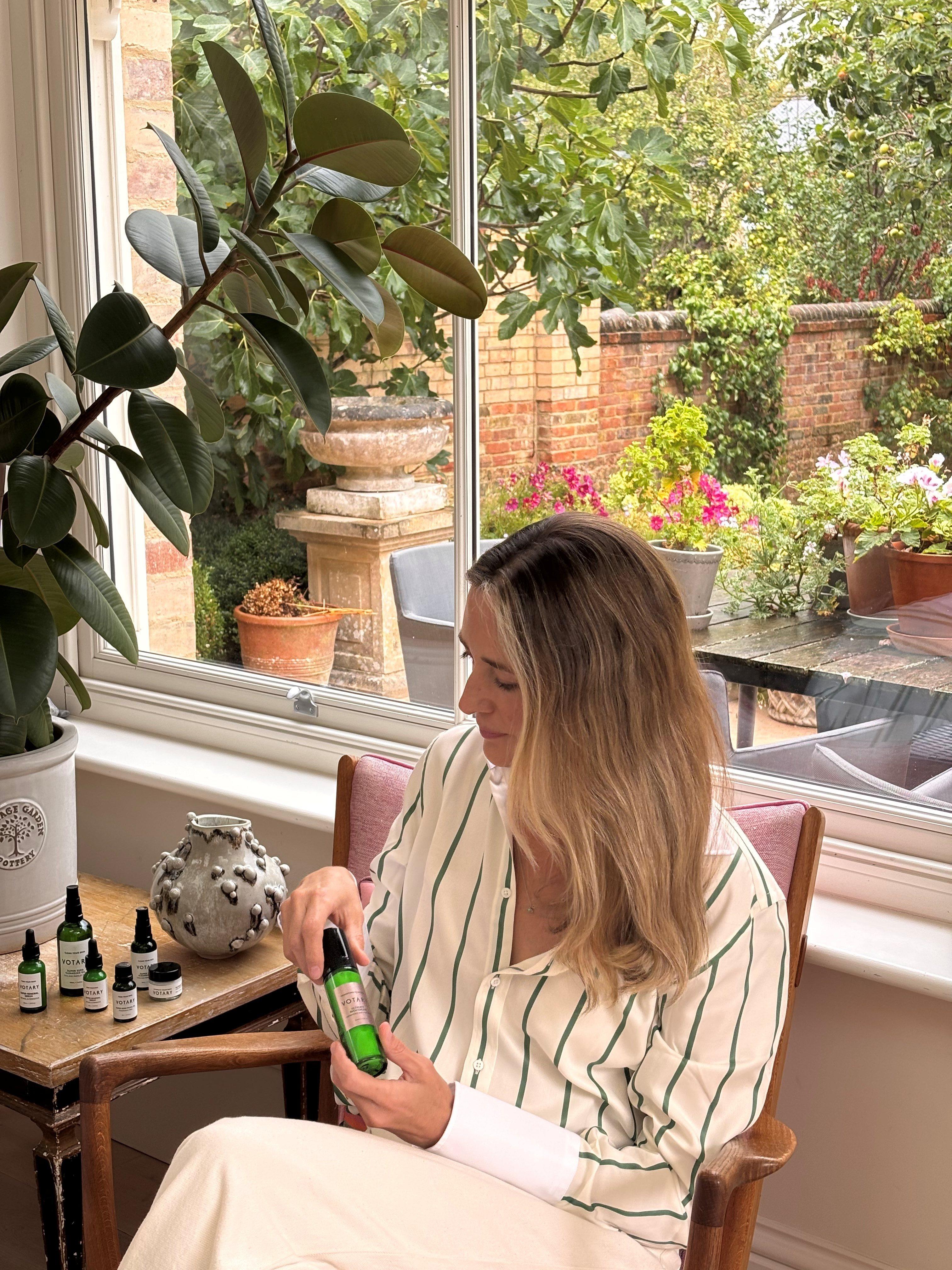 A woman sitting in a chair, holding skincare products, wearing an all-white outfit 