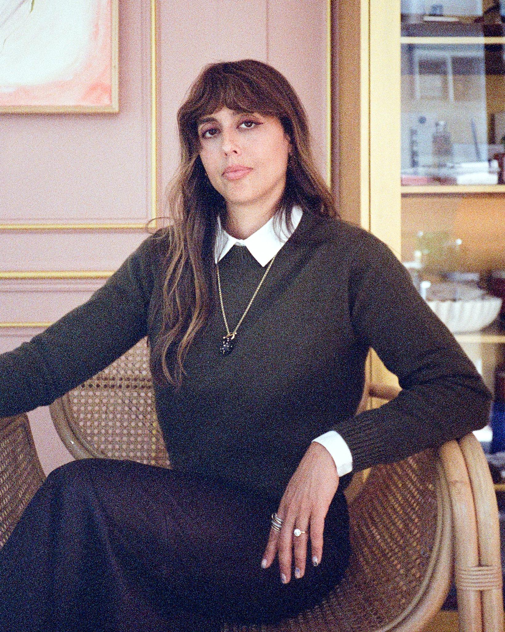 Violette Serrat, the founder of VIOLETTE_FR sits on an ornate chair in a pale pink room. She is wearing a smart jumper over a crisp white shirt with layered gold necklaces