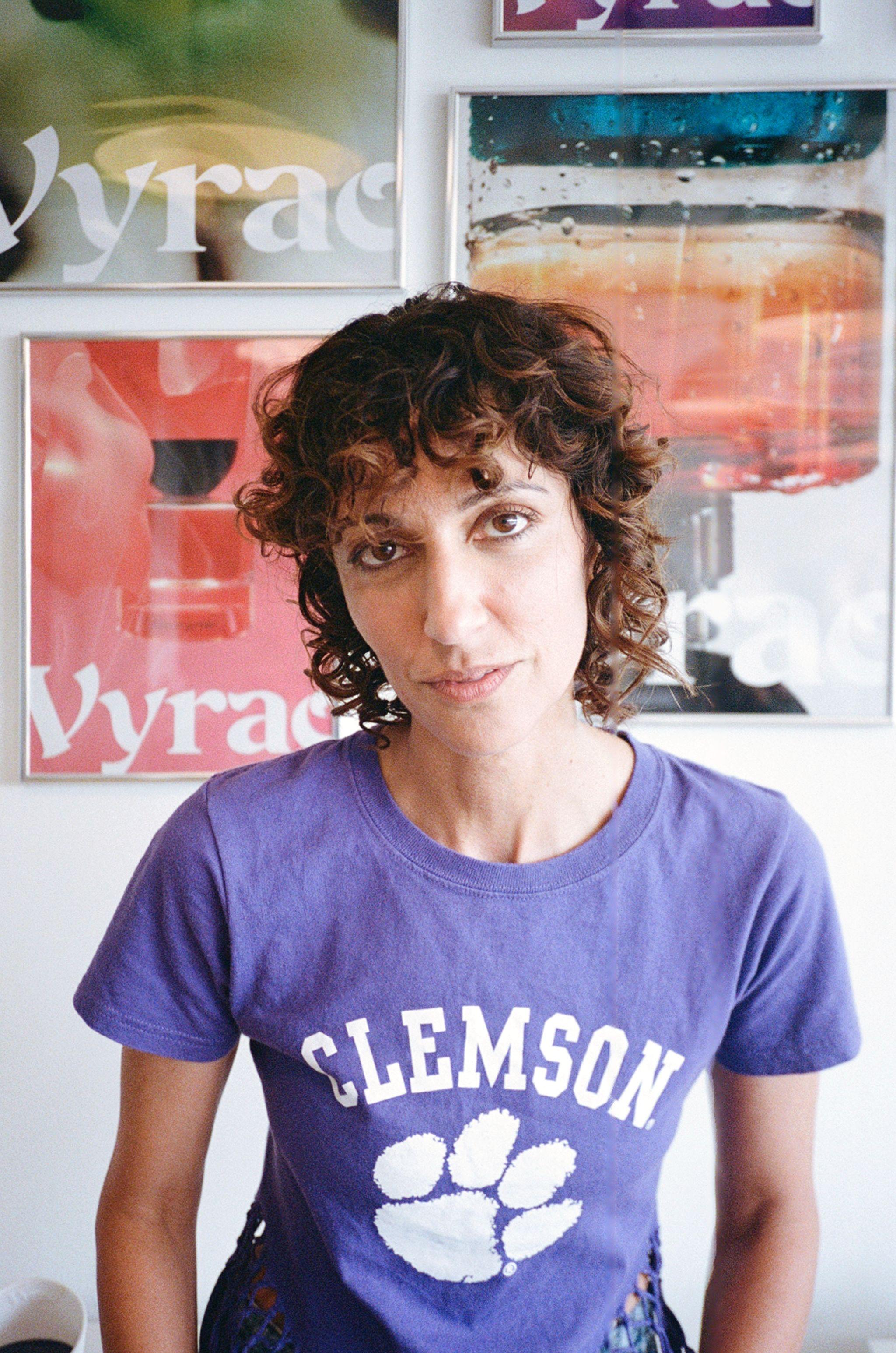 Yasmin Sewell, who has short dark brown curly hair and wears a purple t-shirt, sits in front of a wall of posters from her brand Vyrao, each showing a fragrance bottle in front of vibrant ombre washes of colour 