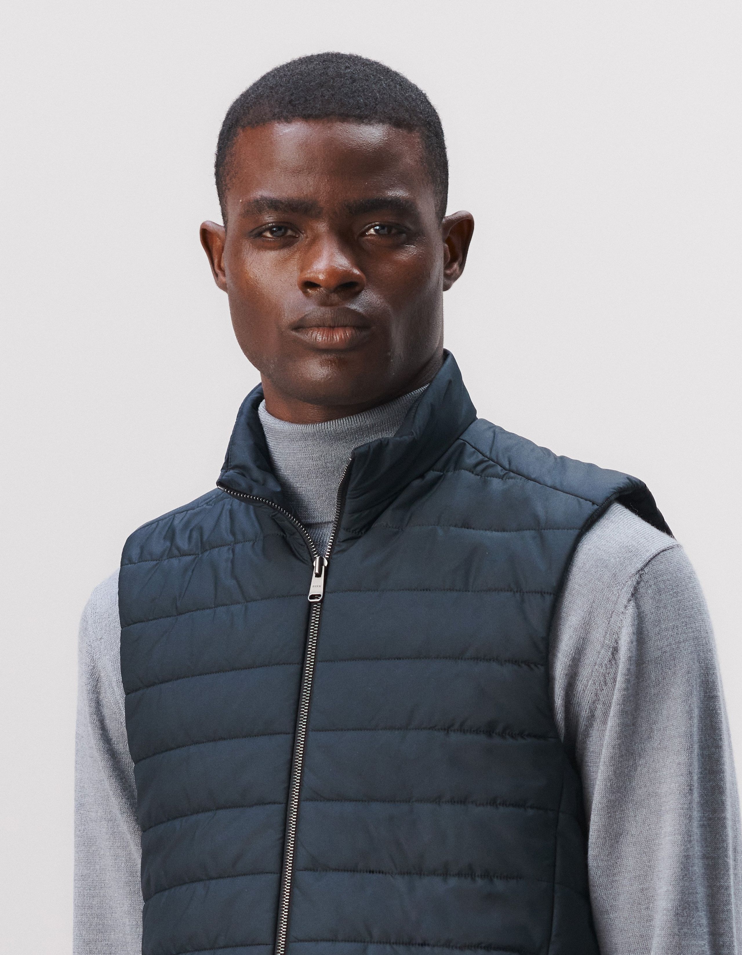 A man wears a grey roll neck sweater with a navy padded gilet