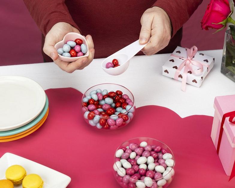 rote, weiße und rosa M&M'S in einer Schale auf einem Tisch mit einer Person, die sie in einen herzförmigen Behälter schöpft