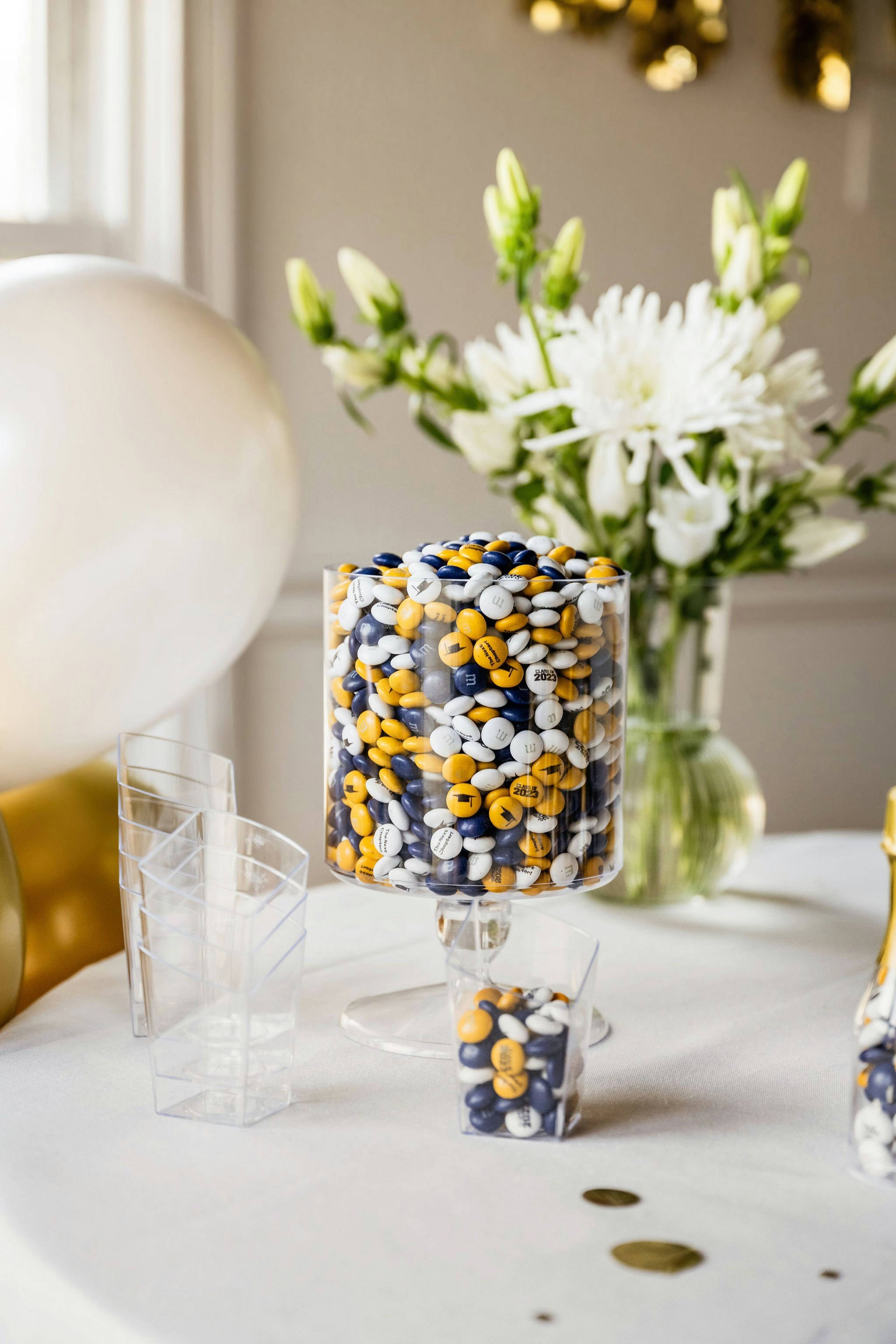 Clear dispenser filled with M&M'S chocolate candies in gold, white, and navy blue with 2023 printed on some candies.