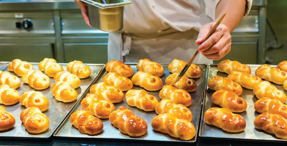 Baker glazing fresh braided rolls on baking trays with a pastry brush.