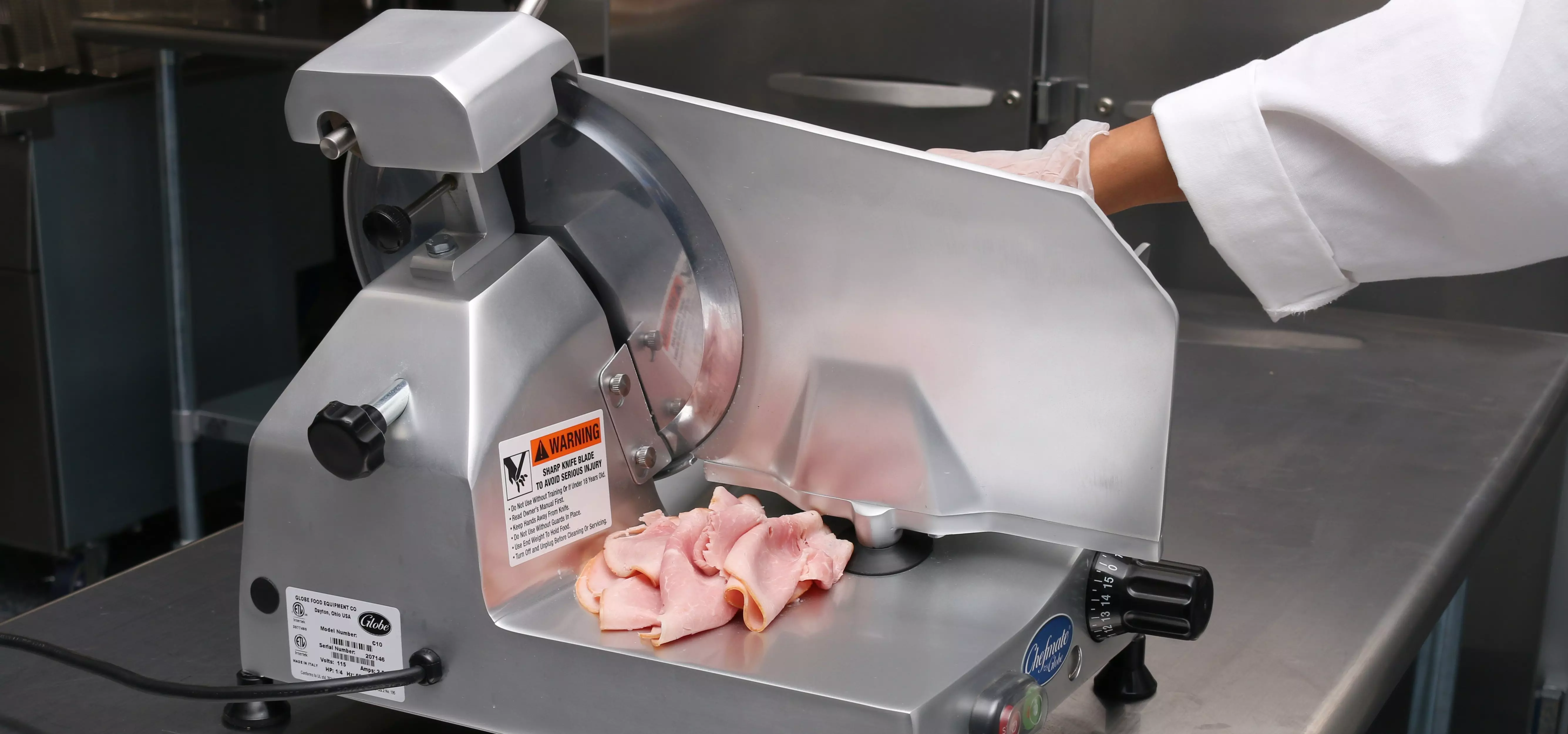 Globe deli meat slicer with sliced ham on the tray and a gloved hand operating it.