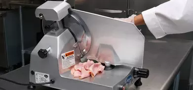 Globe deli meat slicer with sliced ham on the tray and a gloved hand operating it.
