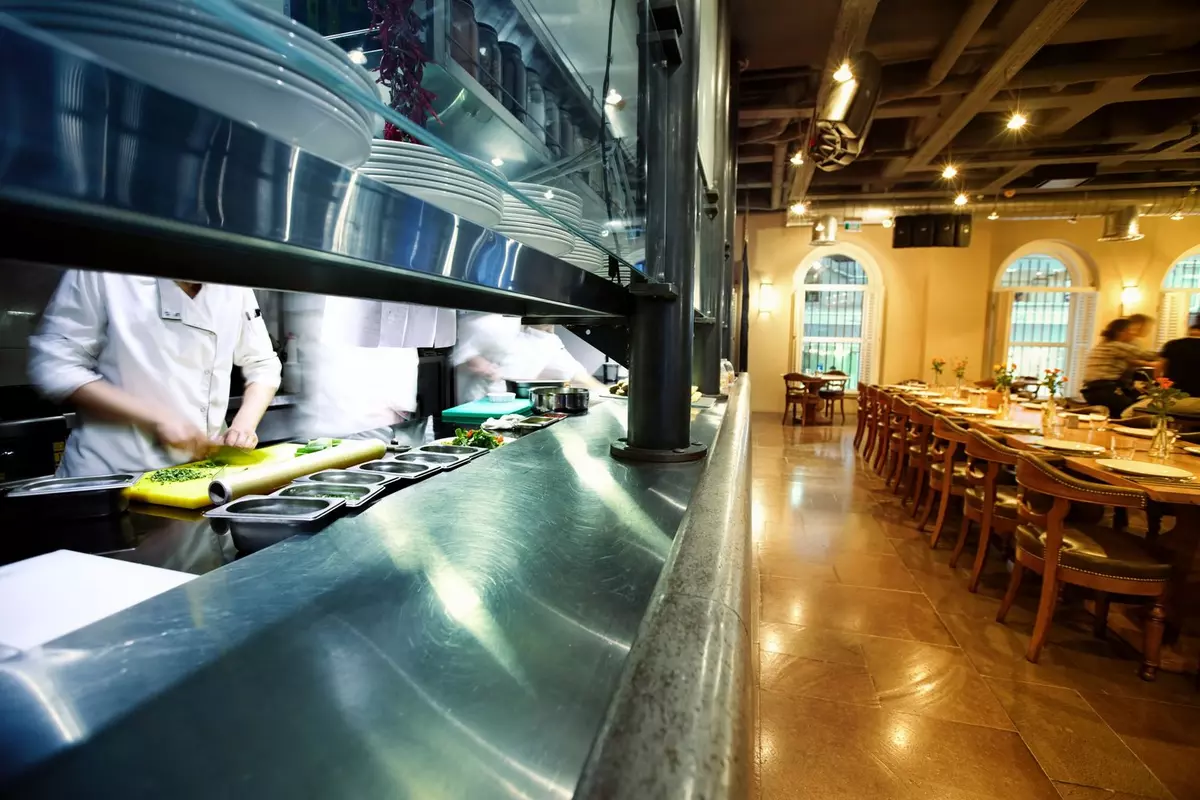 Chefs preparing food at a restaurant pass, with a dining area and set tables beside it.