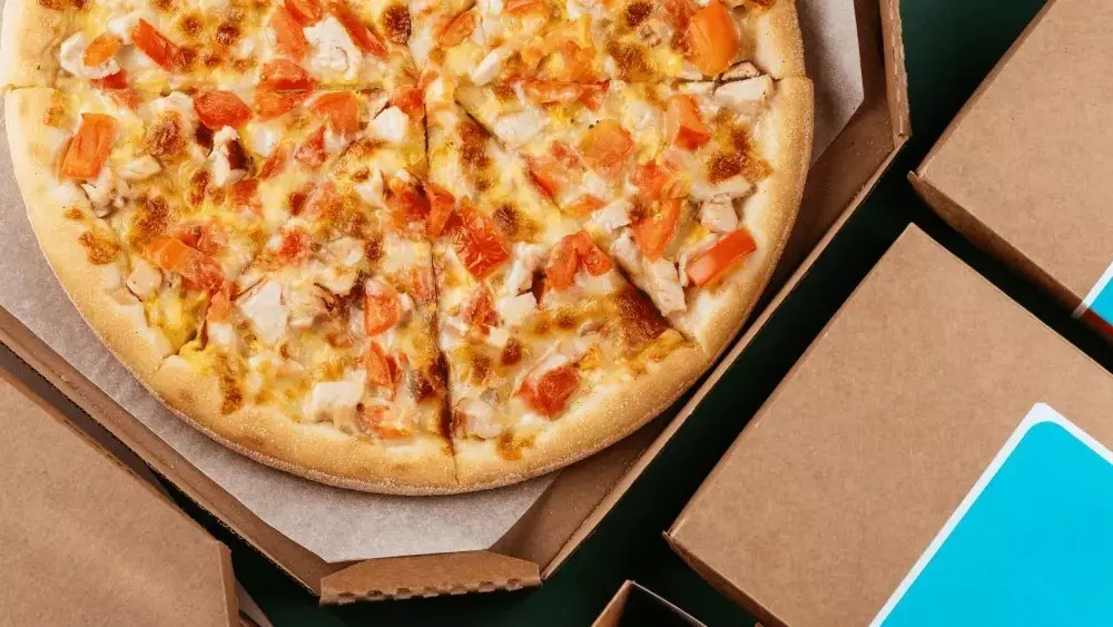 Pizza with cheese, chicken, and tomato in an open box surrounded by closed boxes.