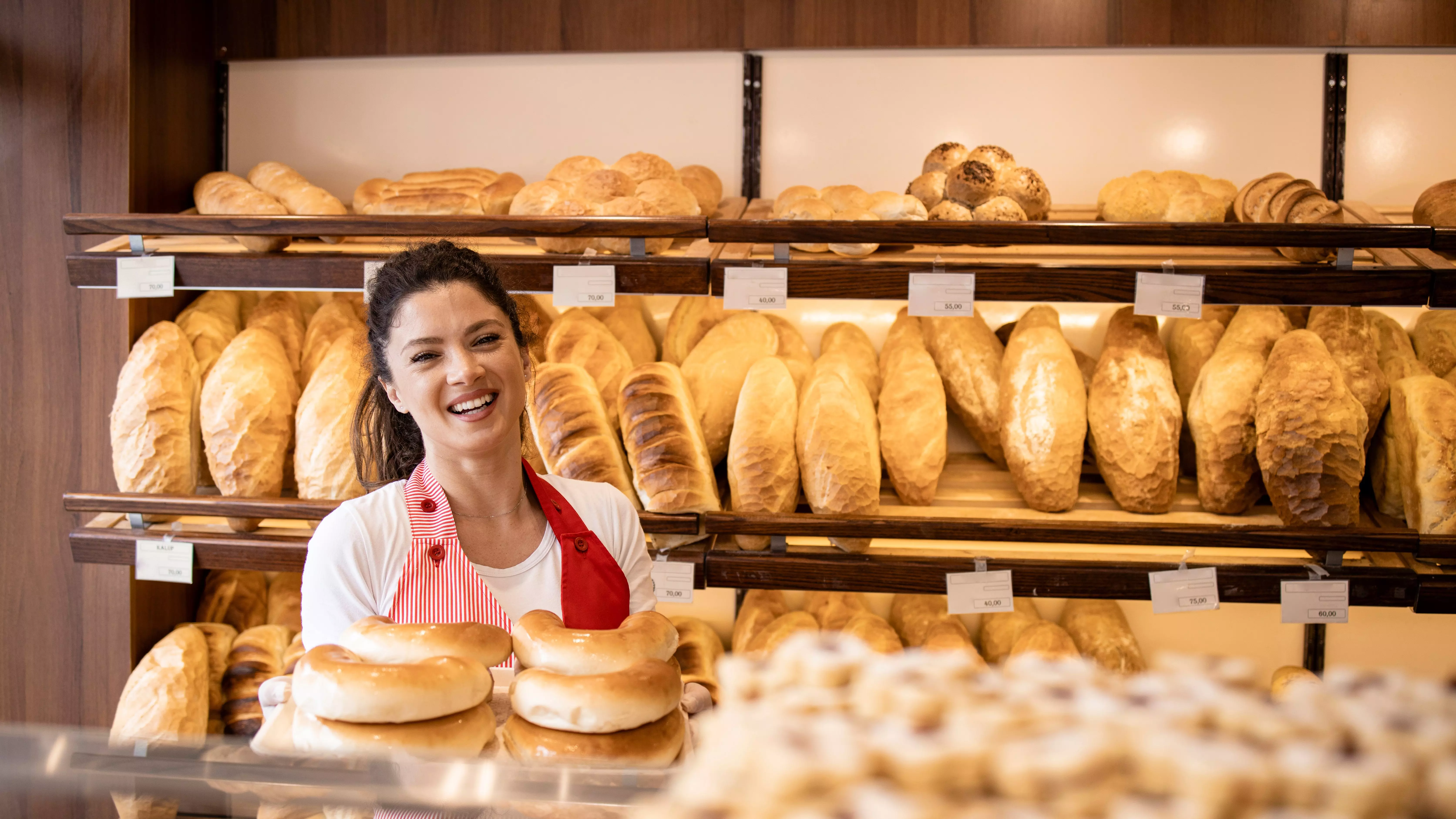 Smiling baker in red apron holding bagels in front of shelves filled with bread.