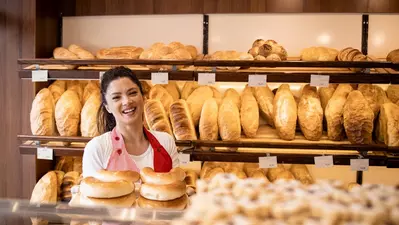 Smiling baker in red apron holding bagels in front of shelves filled with bread.