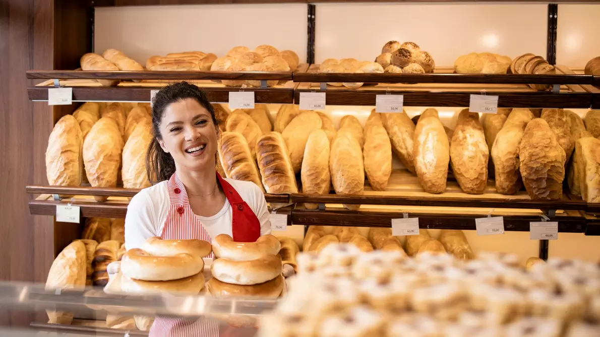 Smiling baker in red apron holding bagels in front of shelves filled with bread.