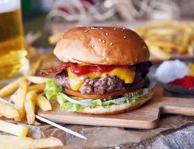 Cheeseburger with lettuce, tomato, onion, bacon, and ketchup on a wooden board, fries and beer nearby.