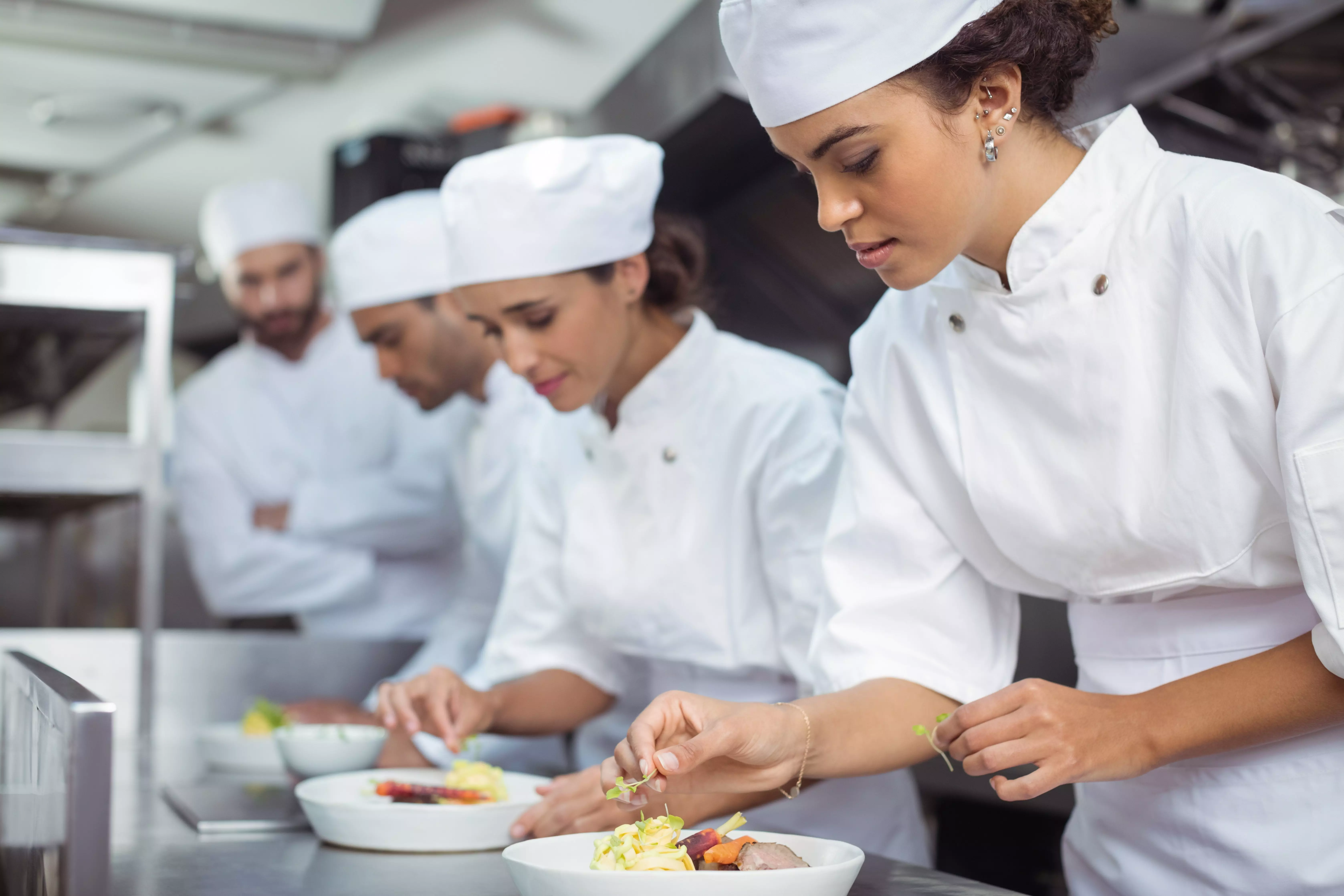 Chef in white uniform garnishing plated dishes in a professional kitchen.