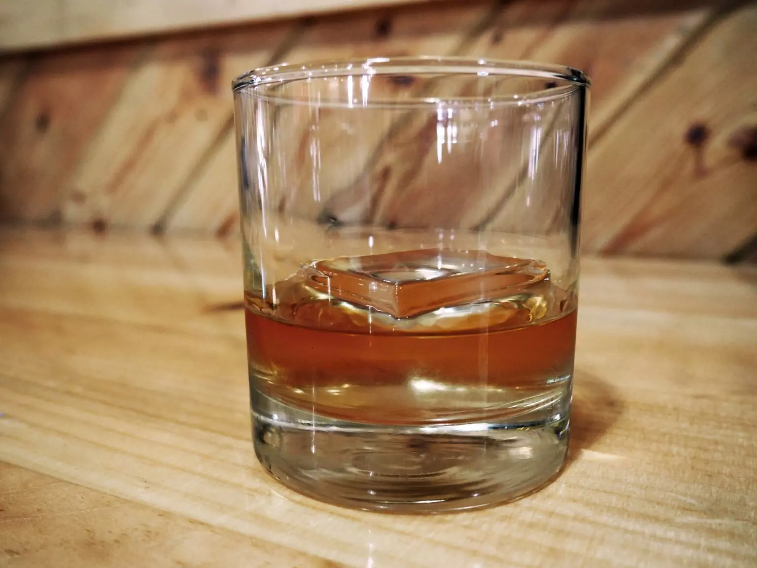 Glass of whiskey with a square ice cube on a wooden surface.