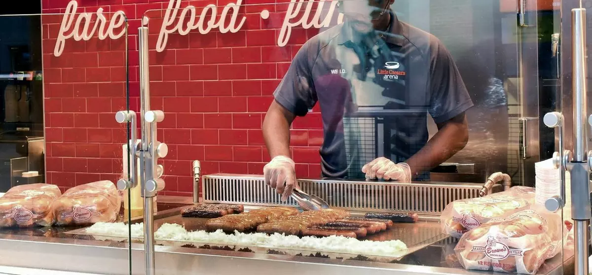 Little Caesars Arena worker grilling burgers and hot dogs behind a glass counter.