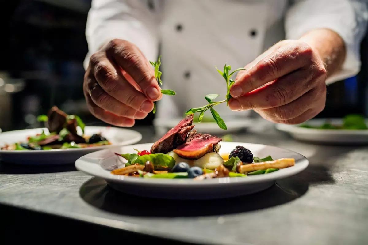 Chef garnishing plated sliced meat with fresh herbs and berries.