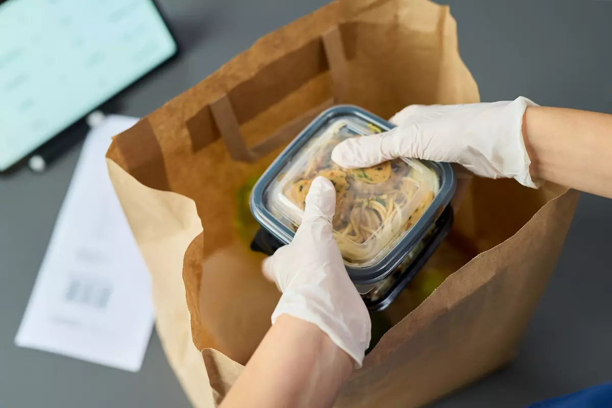 Gloved hands placing a sealed food container into a paper takeout bag.