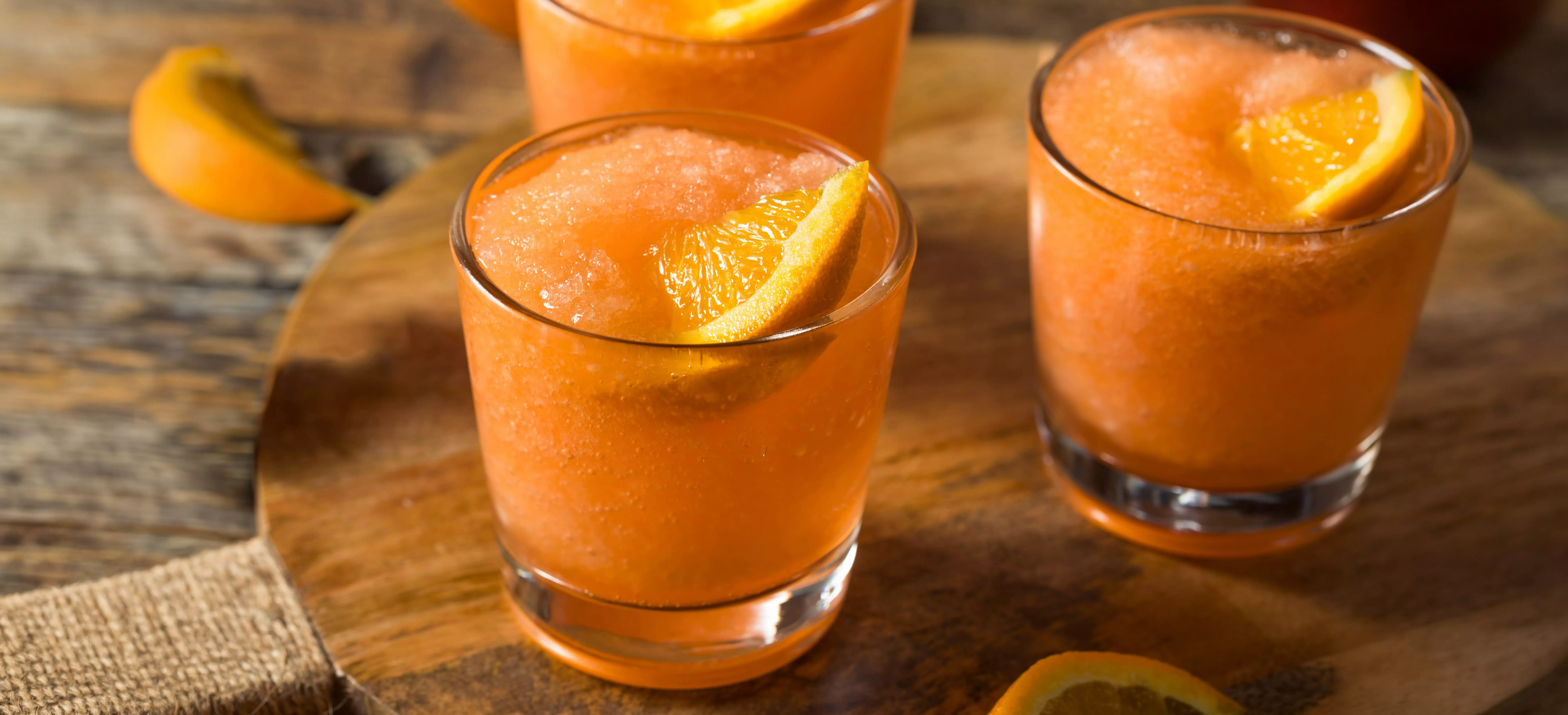 Two glasses of orange frozen drink with orange slice garnish on a wooden board.
